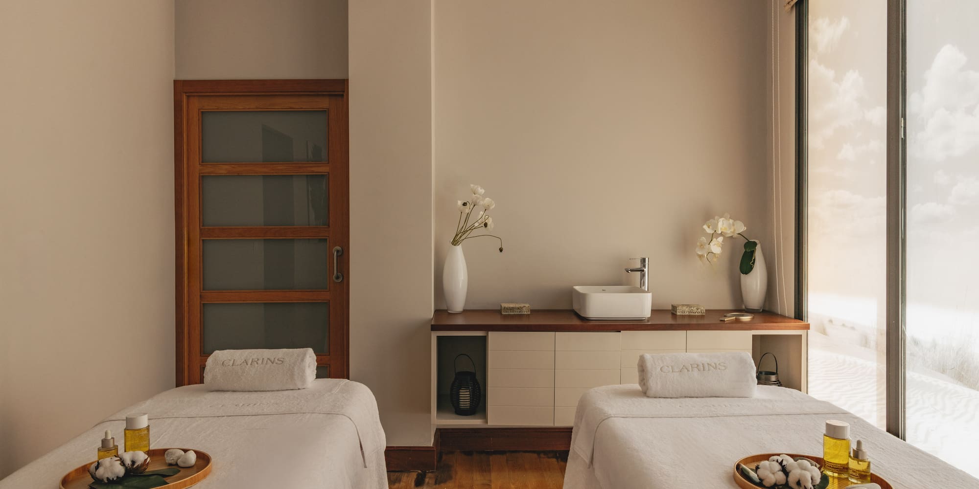 Clarins spa treatment room with two massage beds, oils, and a desert view window.