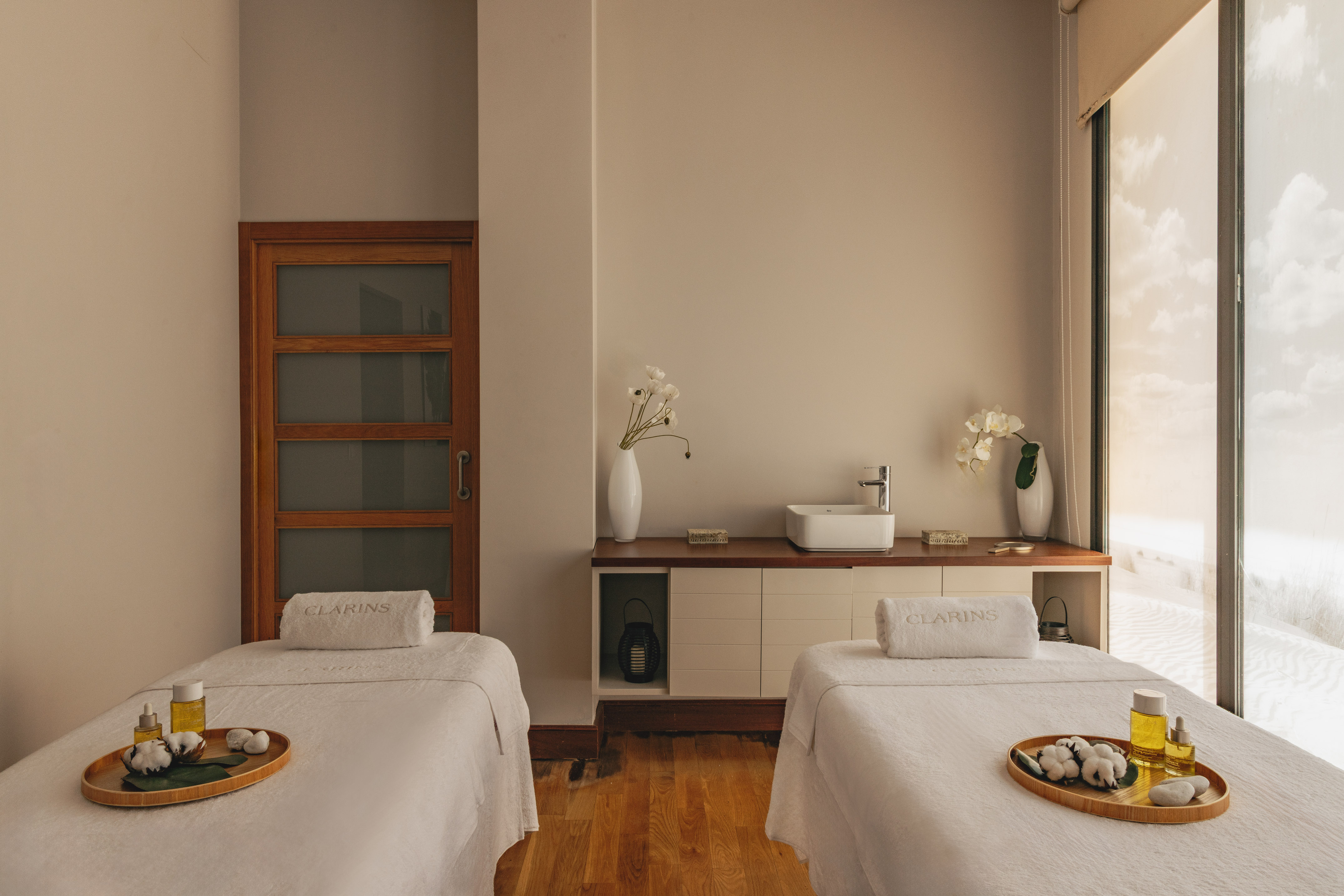 Clarins spa treatment room with two massage beds, oils, and a desert view window.