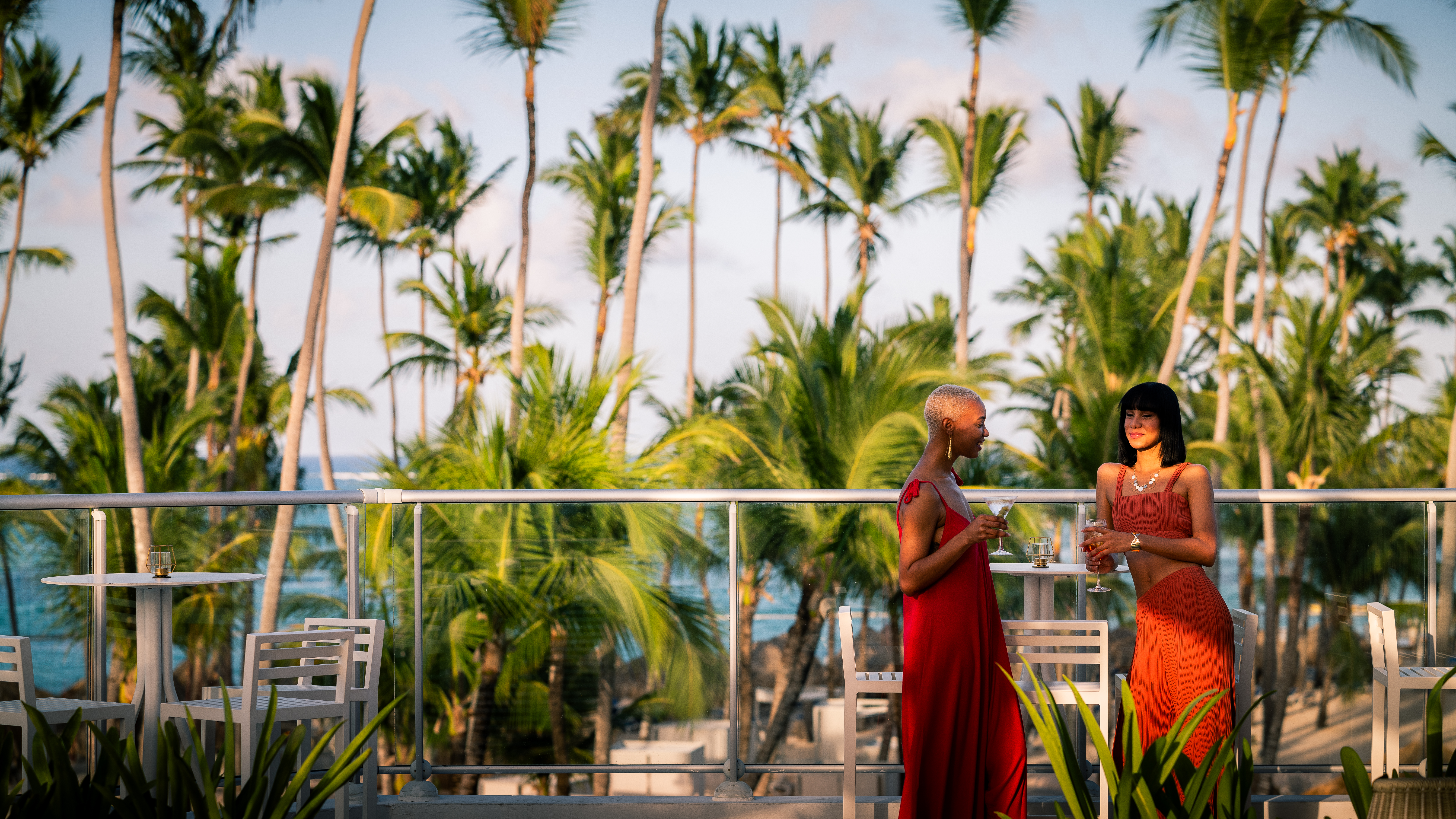 Elegant women with cocktails on tropical resort balcony at sunset.