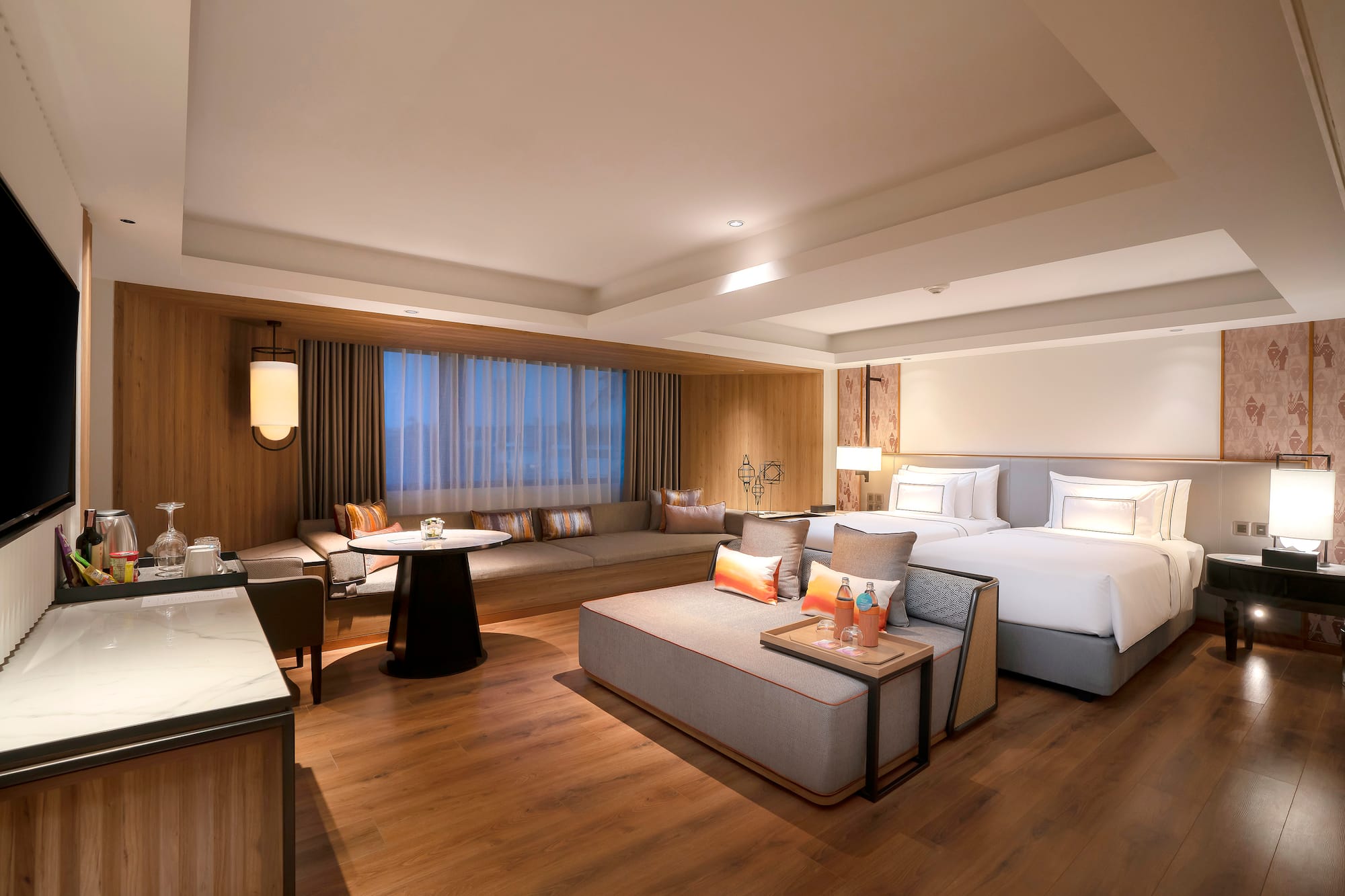Modern hotel suite with two beds, lounge area, wood floors, and large window.