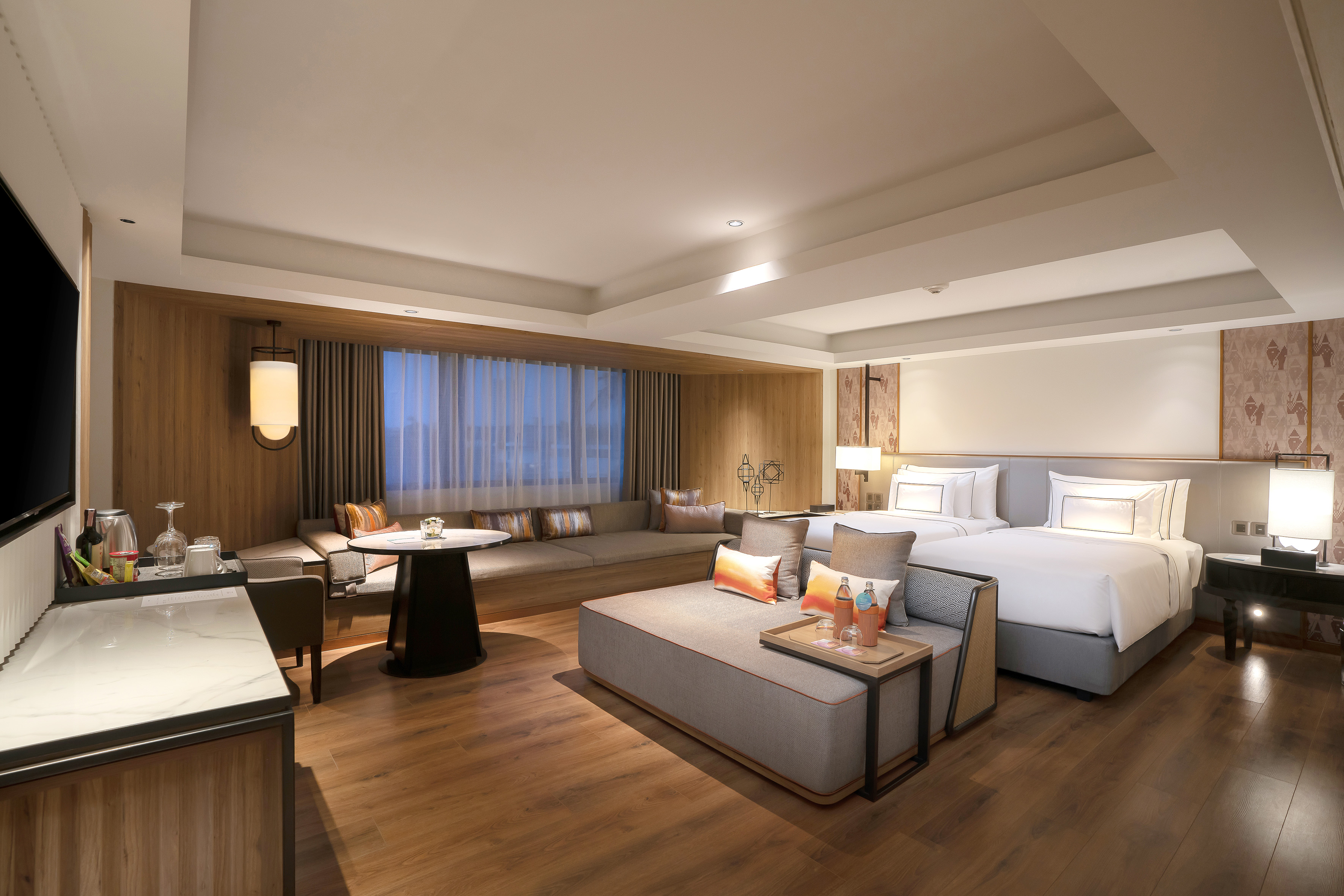 Modern hotel suite with two beds, lounge area, wood floors, and large window.