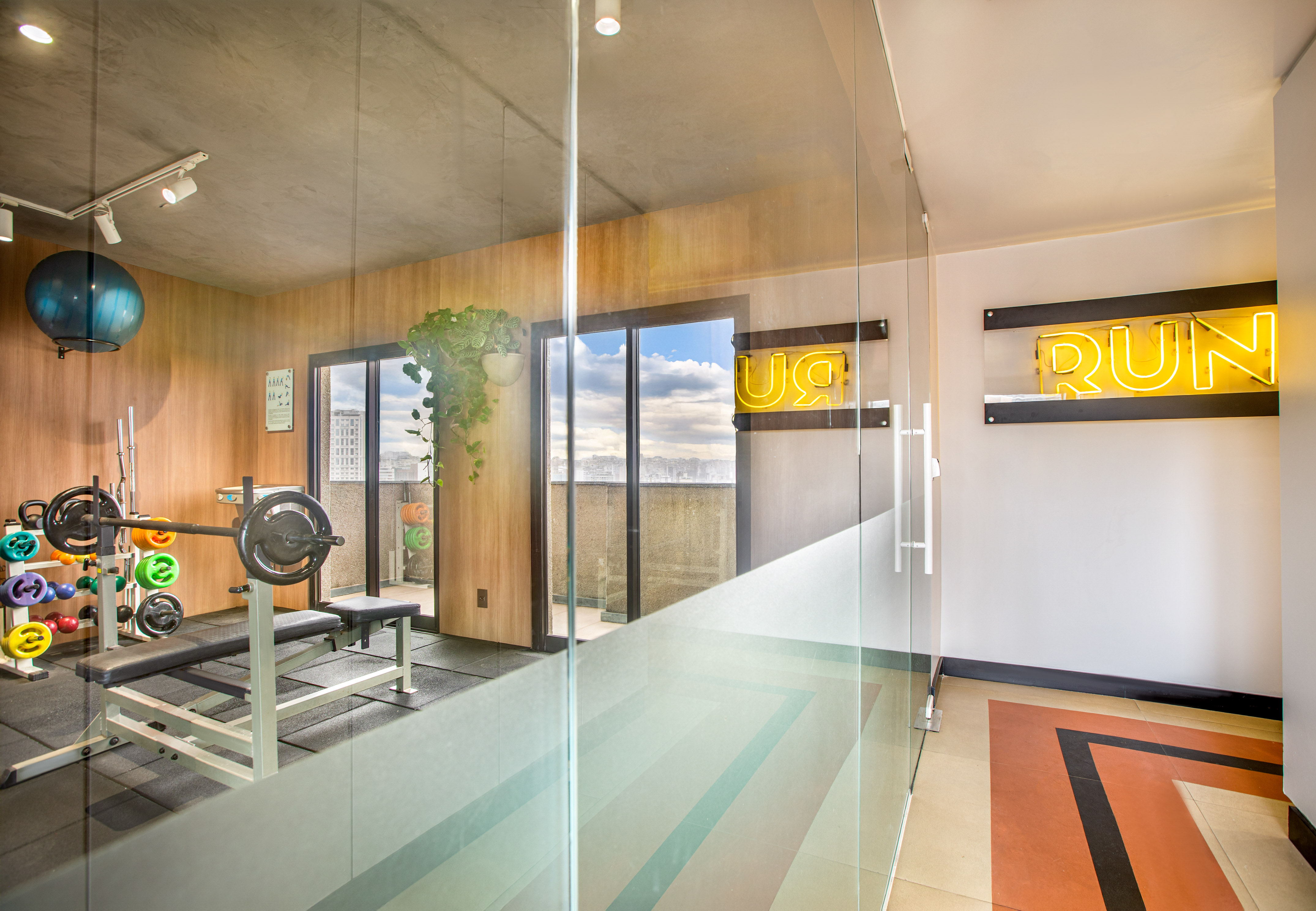Modern home gym with bench press, weights, "RUN" neon sign, and city view.