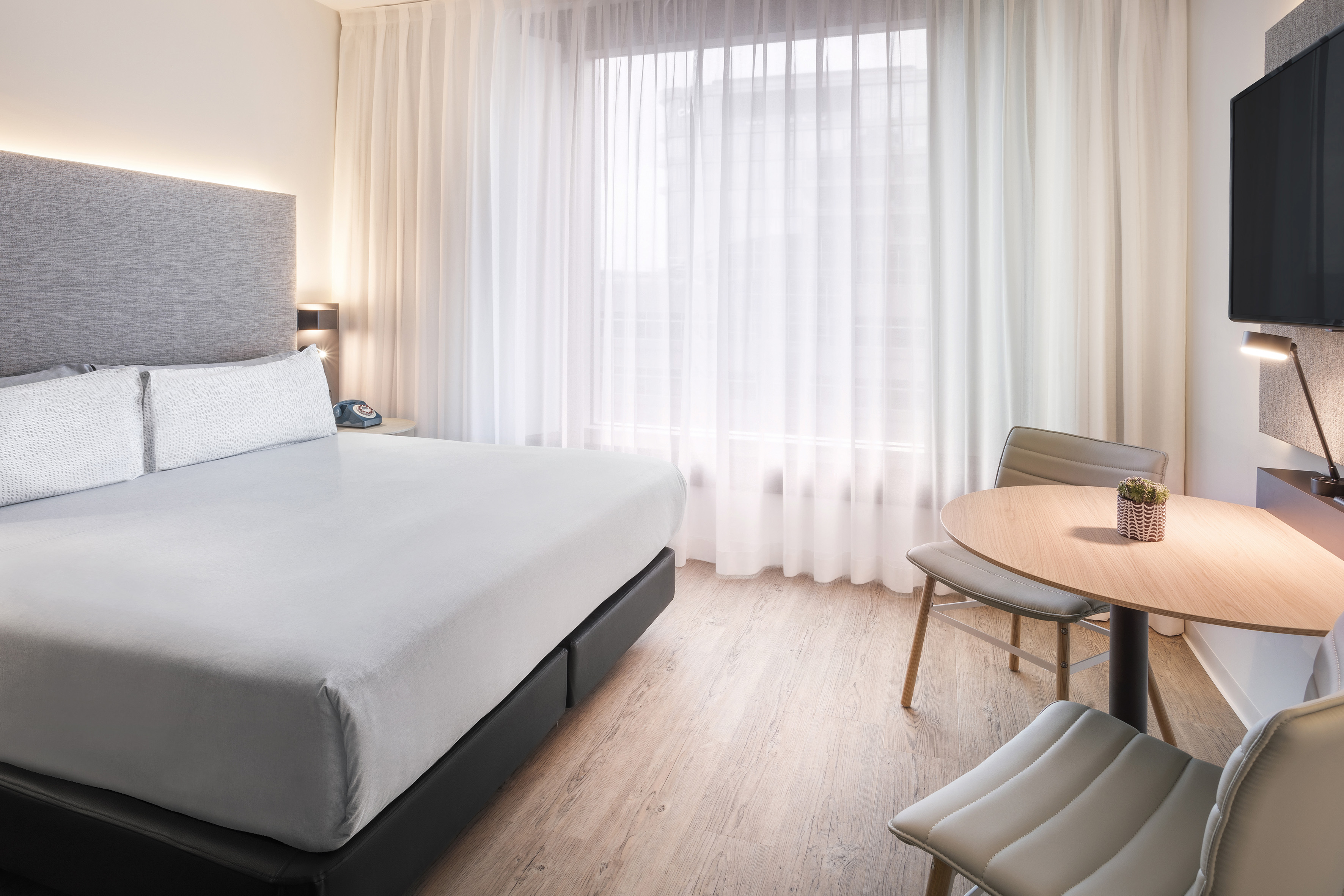 Modern hotel bedroom with king bed, sheer curtains, small table, and TV.