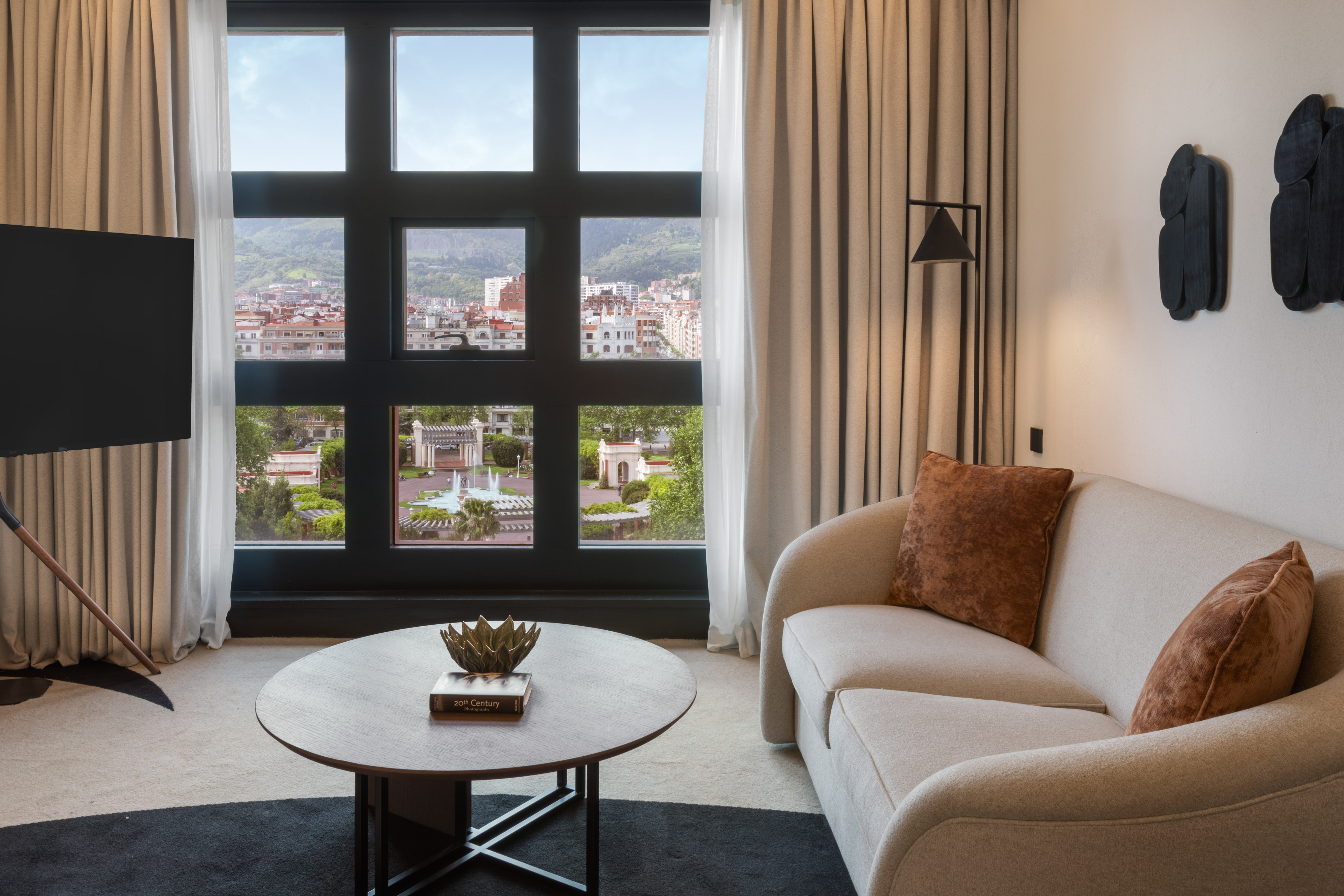 Modern living room with city and park view, beige sofa, coffee table.