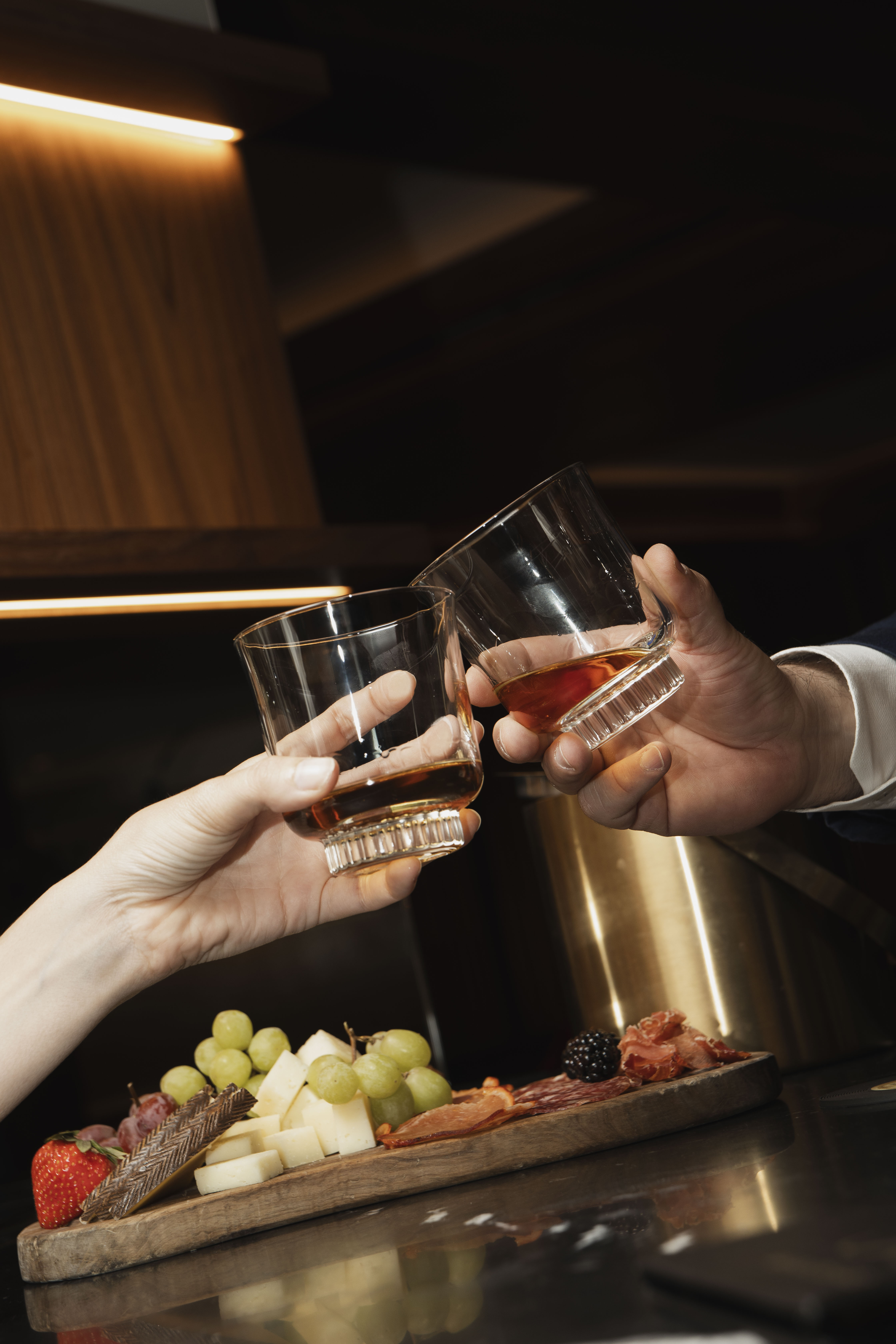 Couple toasting whiskey glasses over a cheese and charcuterie board.