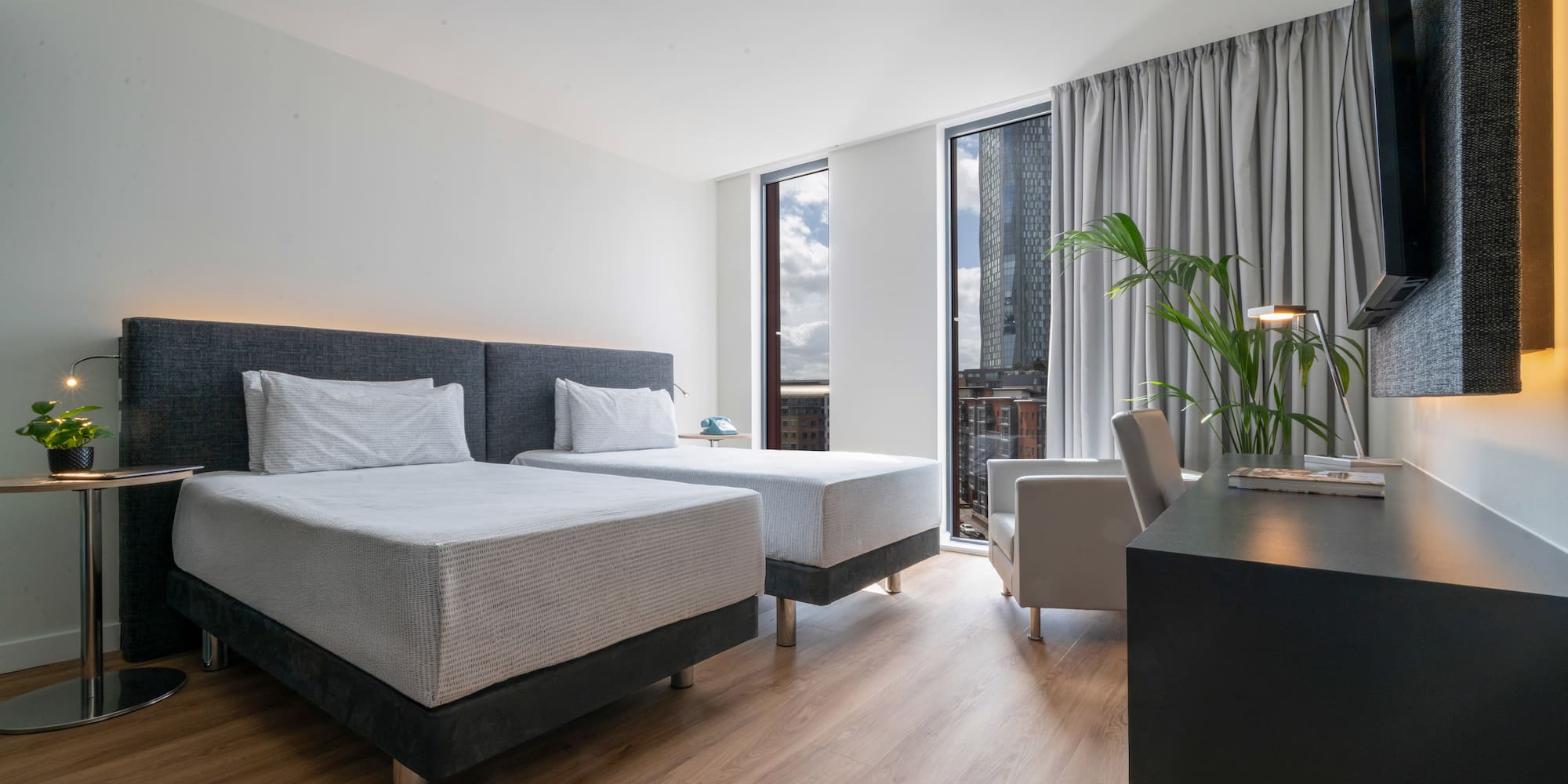 Modern hotel twin room with city view, wooden floor, and work desk.