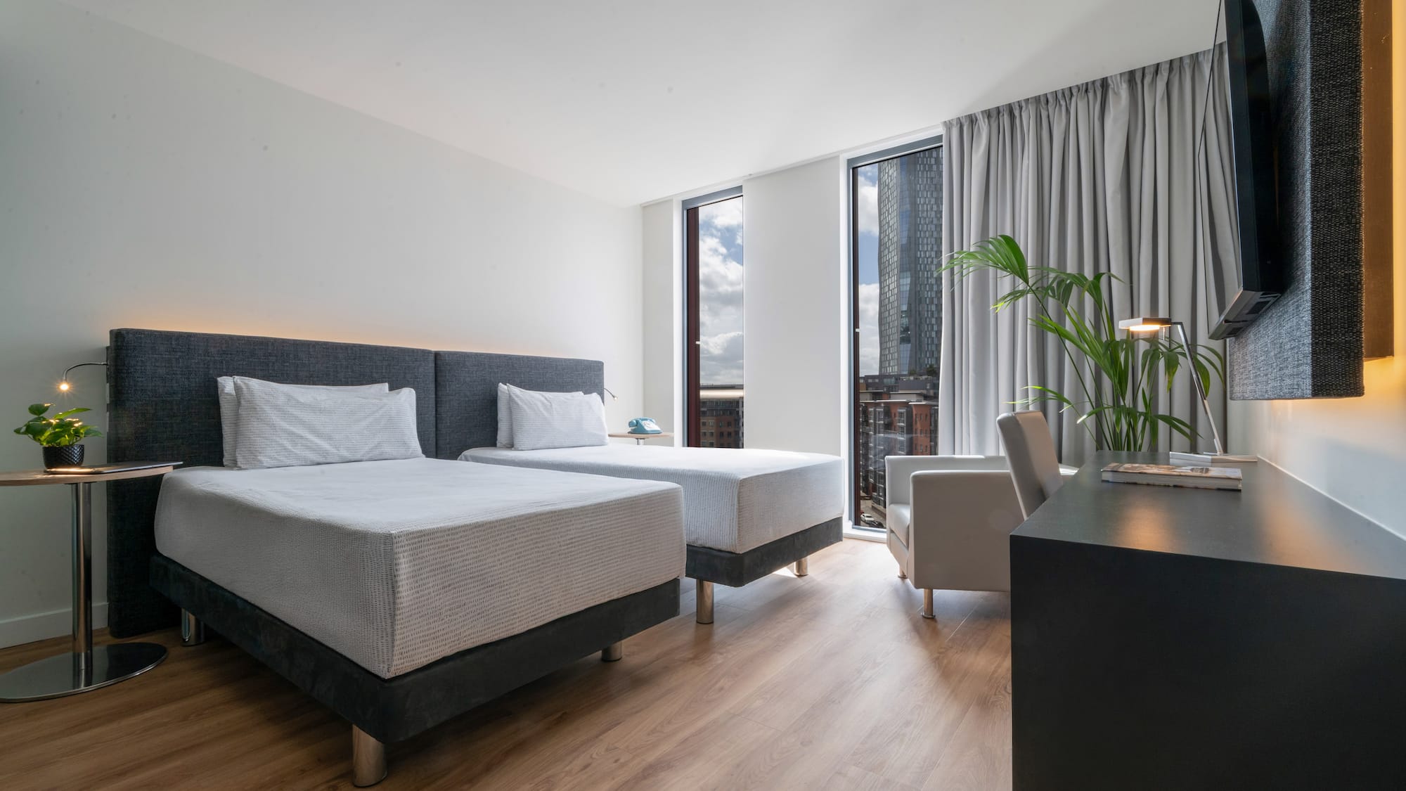 Modern hotel twin room with city view, wooden floor, and work desk.