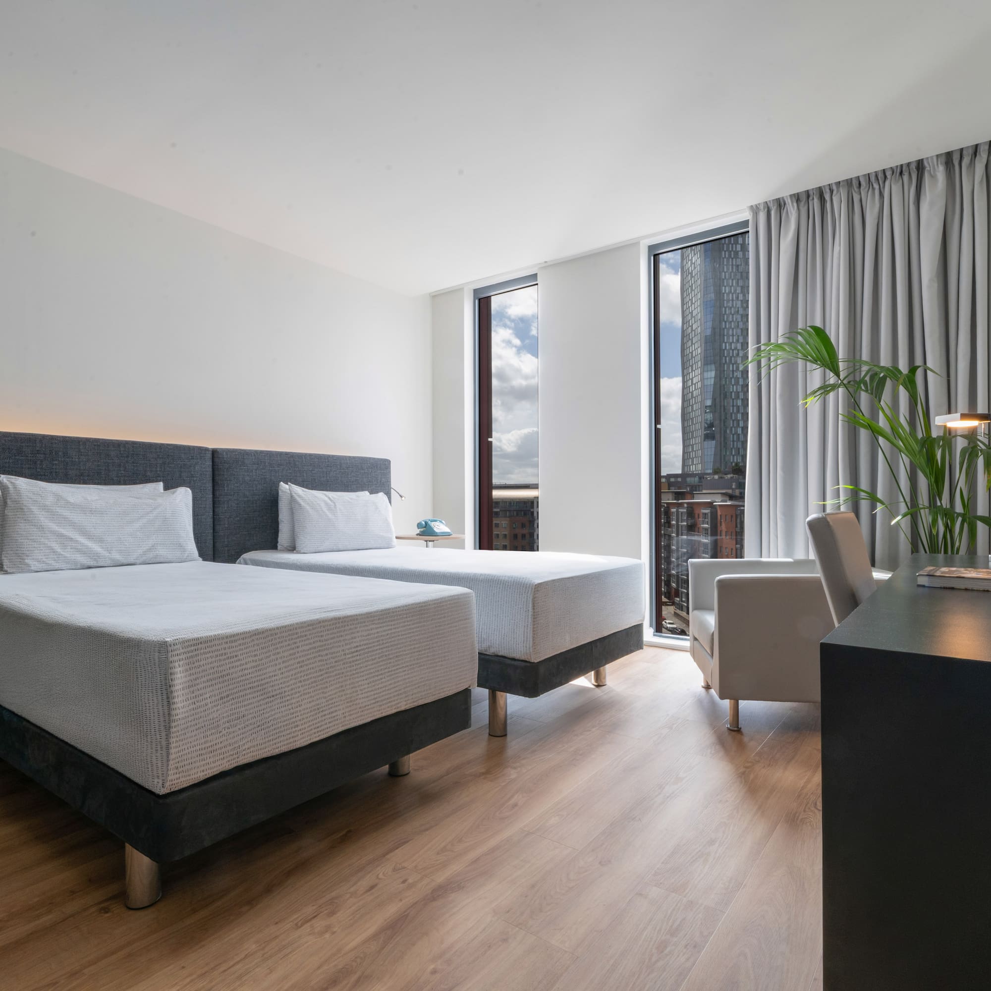 Modern hotel twin room with city view, wooden floor, and work desk.