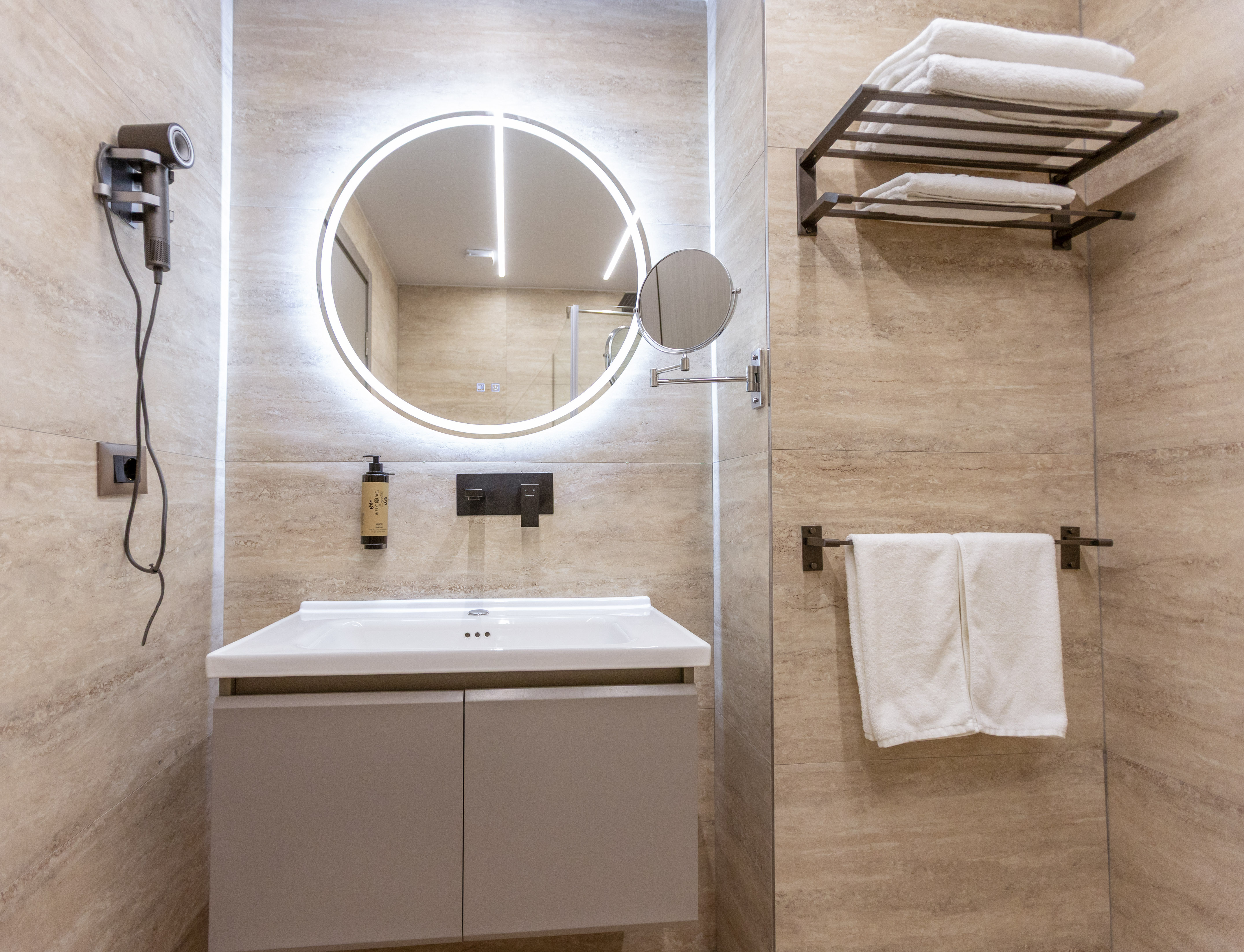 Modern bathroom vanity, round LED mirror, hairdryer, white towels, stone tiles.