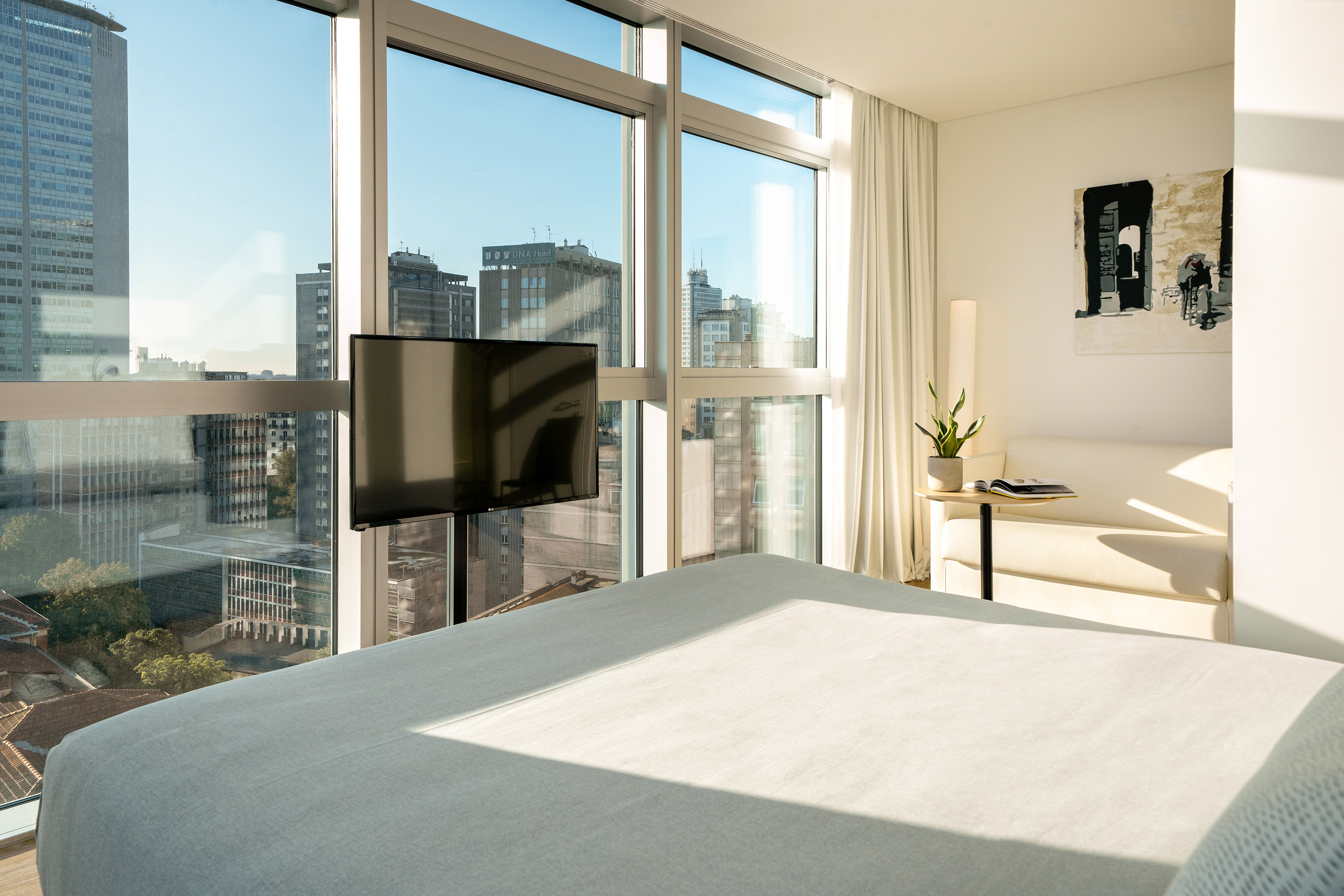 Modern hotel bedroom with city view, large windows, TV, and white bed.