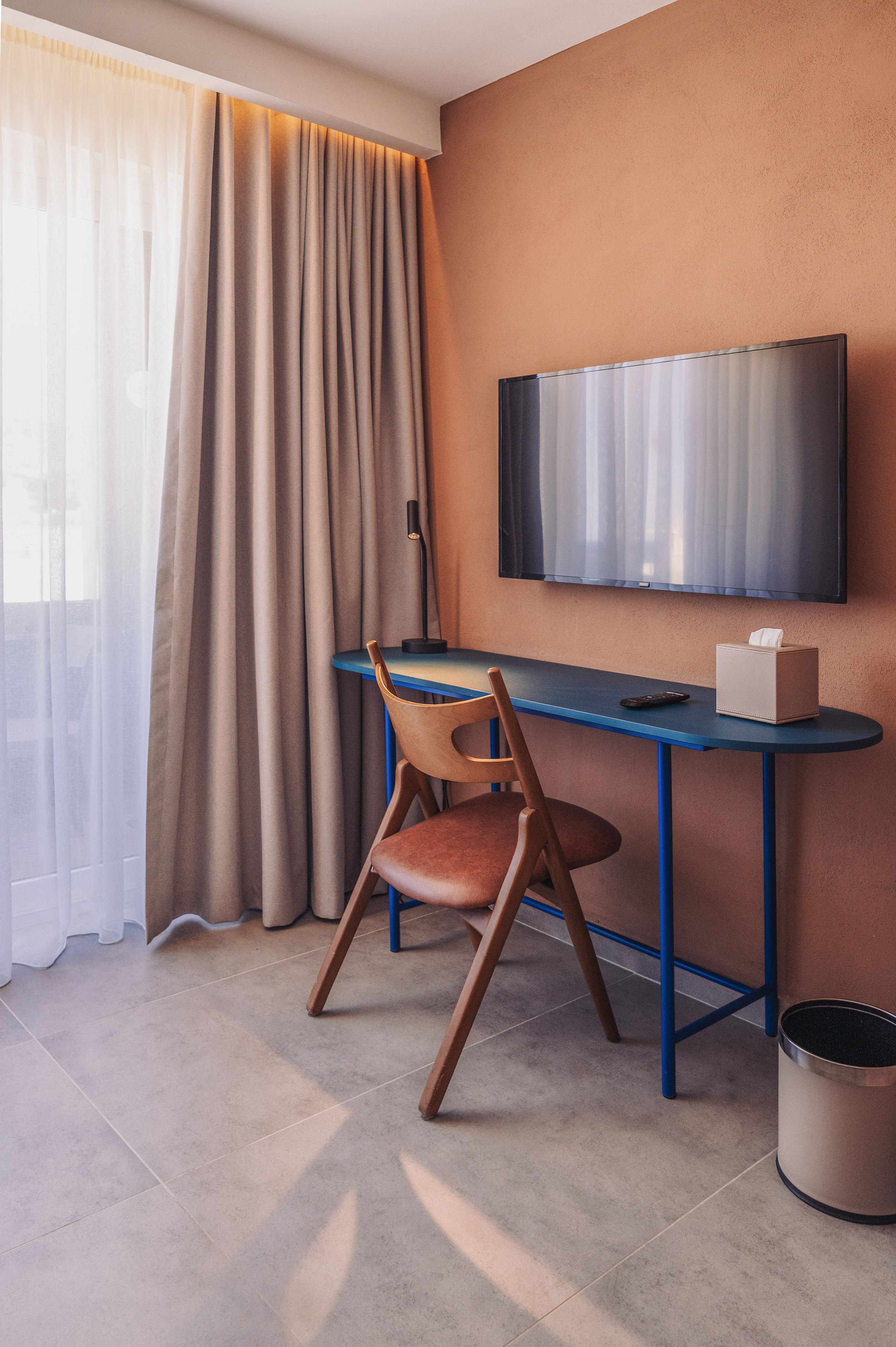 Contemporary hotel workspace with blue desk, wooden chair, TV, and orange wall.