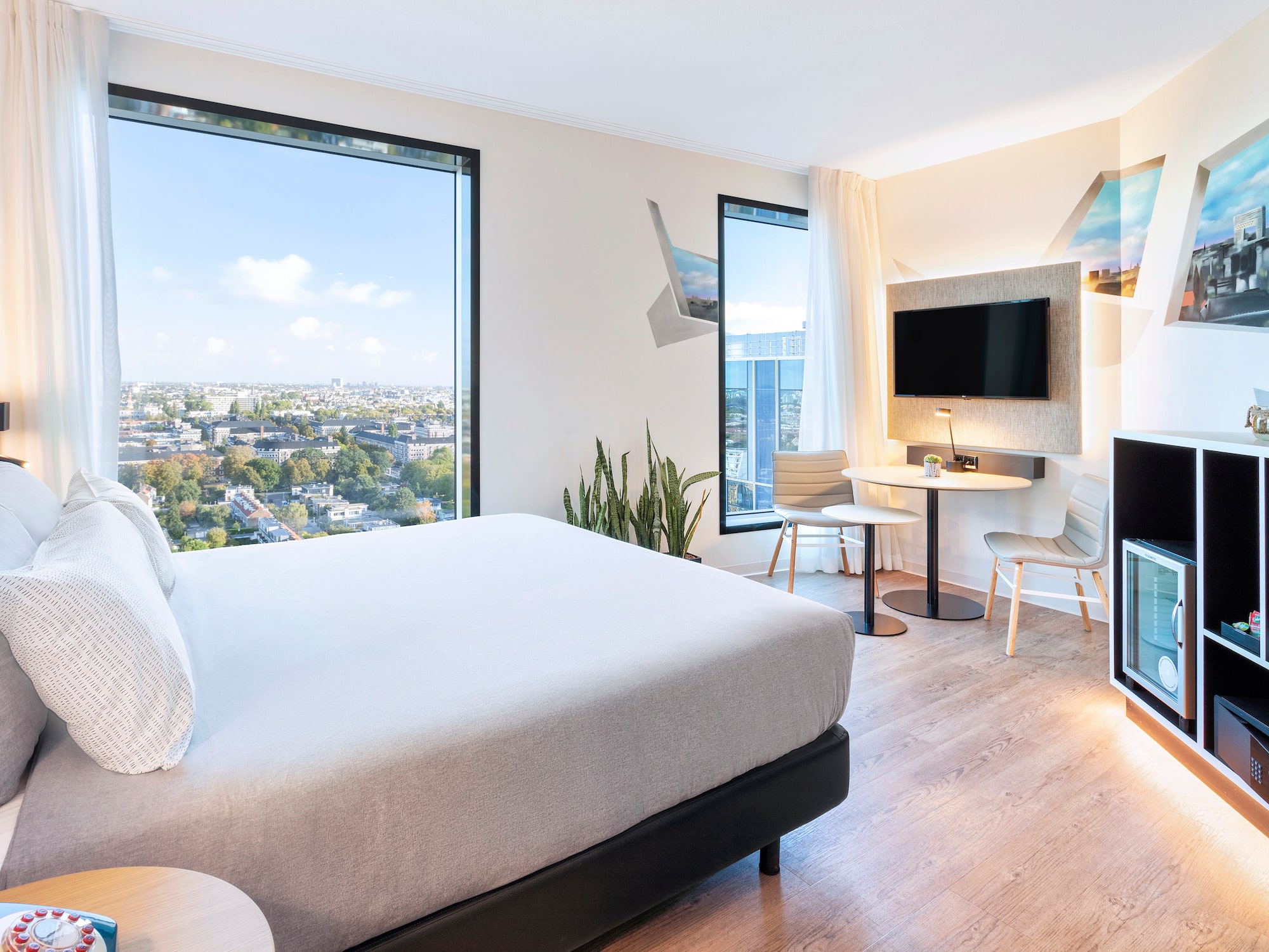 Modern hotel room with city view, large bed, TV, and chic furnishings.