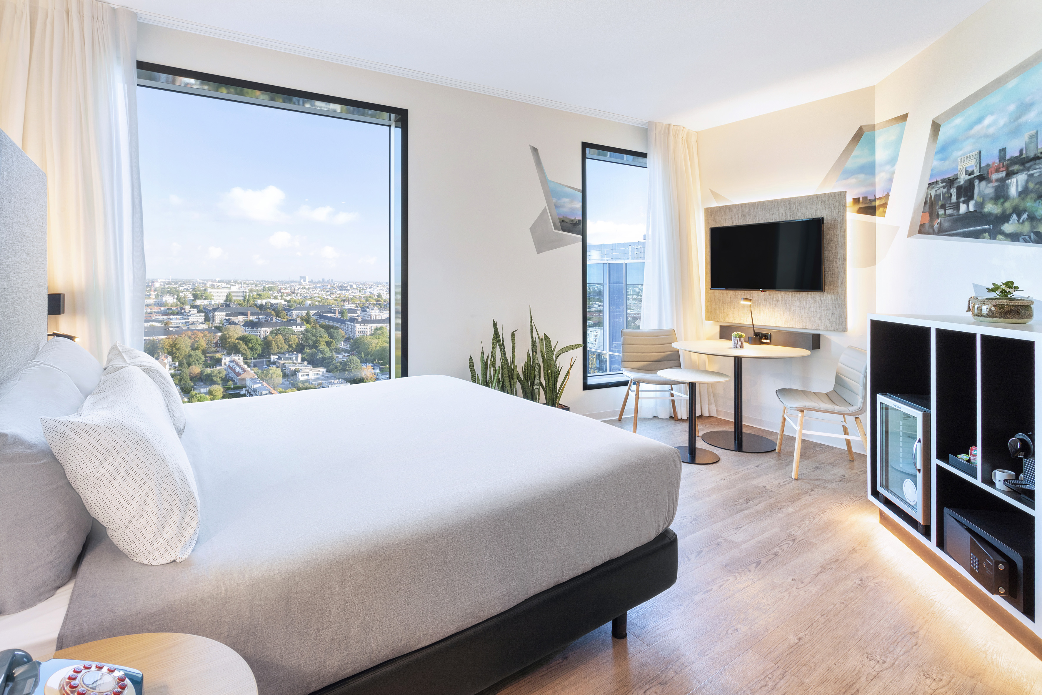 Modern hotel room with city view, large bed, TV, and chic furnishings.