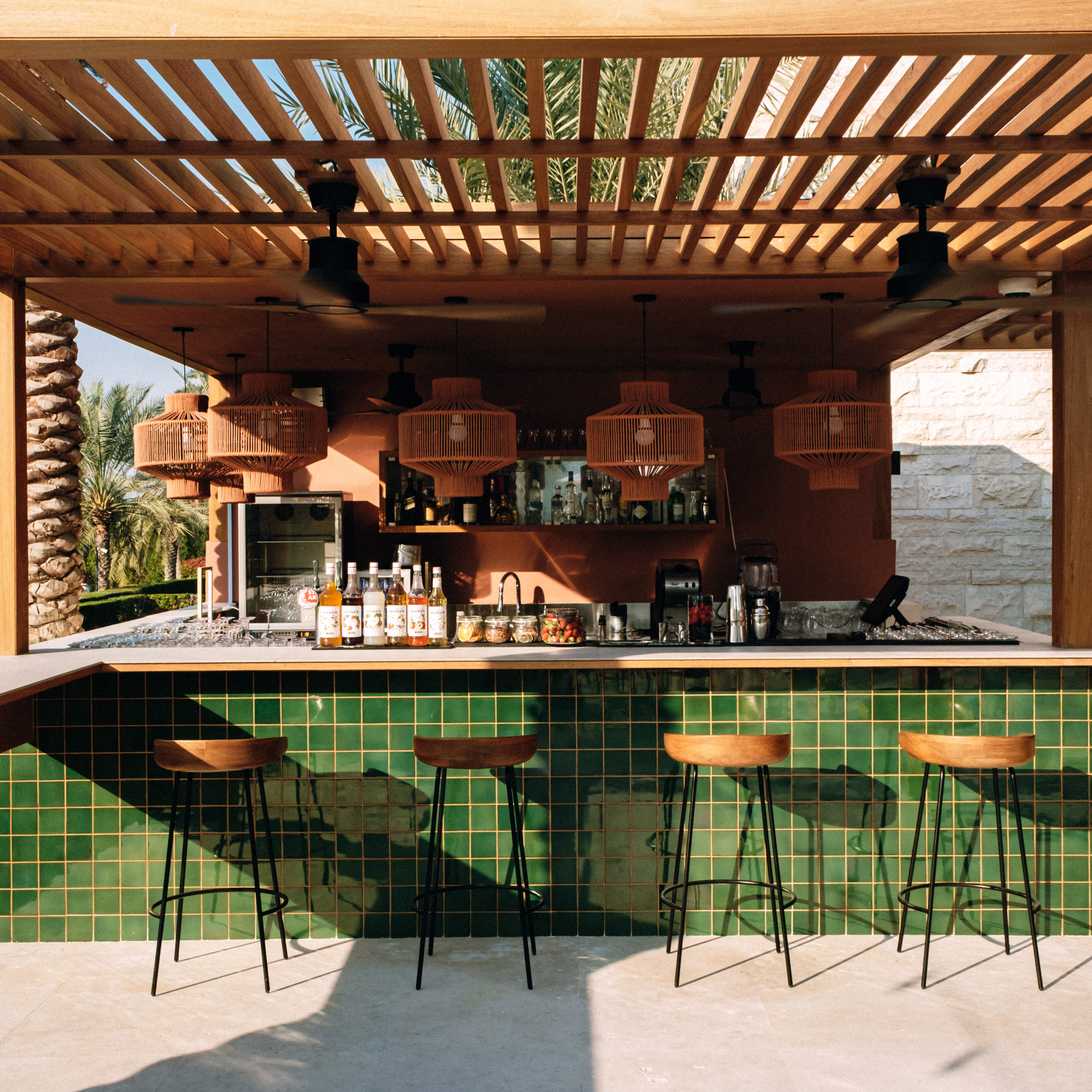 Tropical outdoor bar with green tiled counter, wooden stools, and pergola.