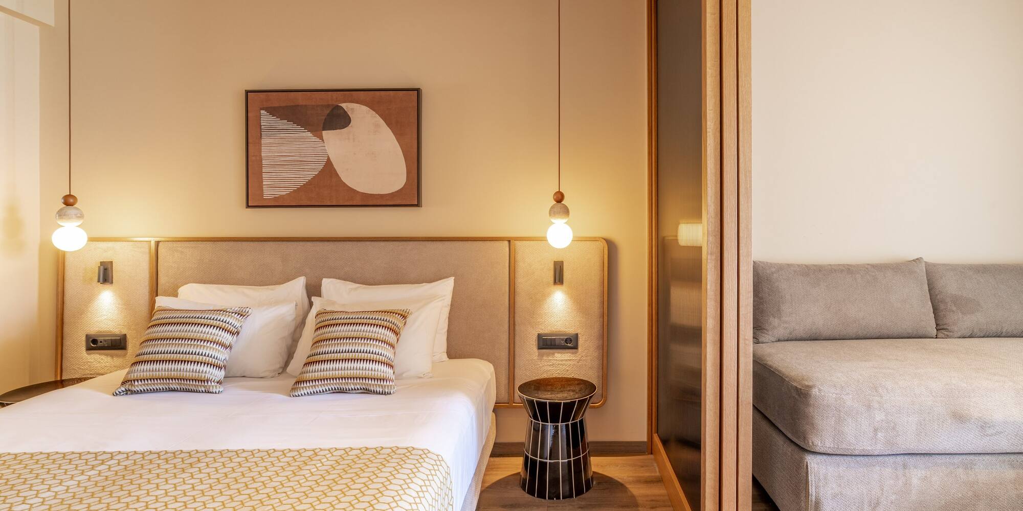 Modern hotel bedroom with large bed, abstract art, pendant light, and gray sofa.
