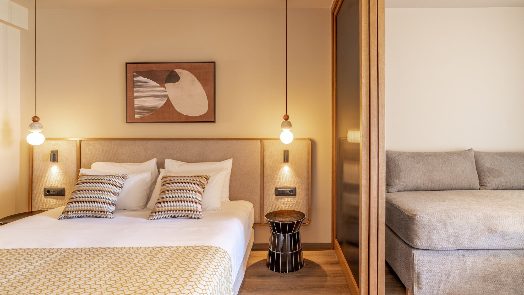 Modern hotel bedroom with large bed, abstract art, pendant light, and gray sofa.