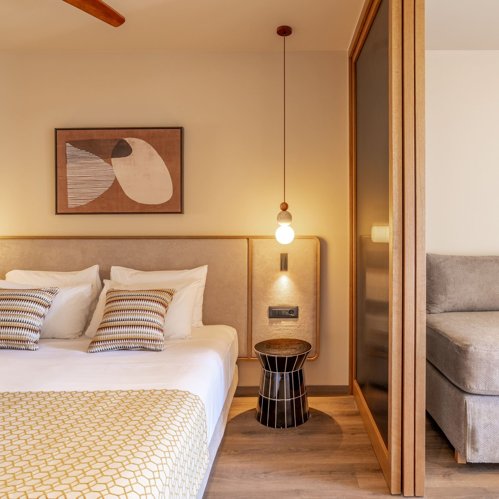 Modern hotel bedroom with large bed, abstract art, pendant light, and gray sofa.