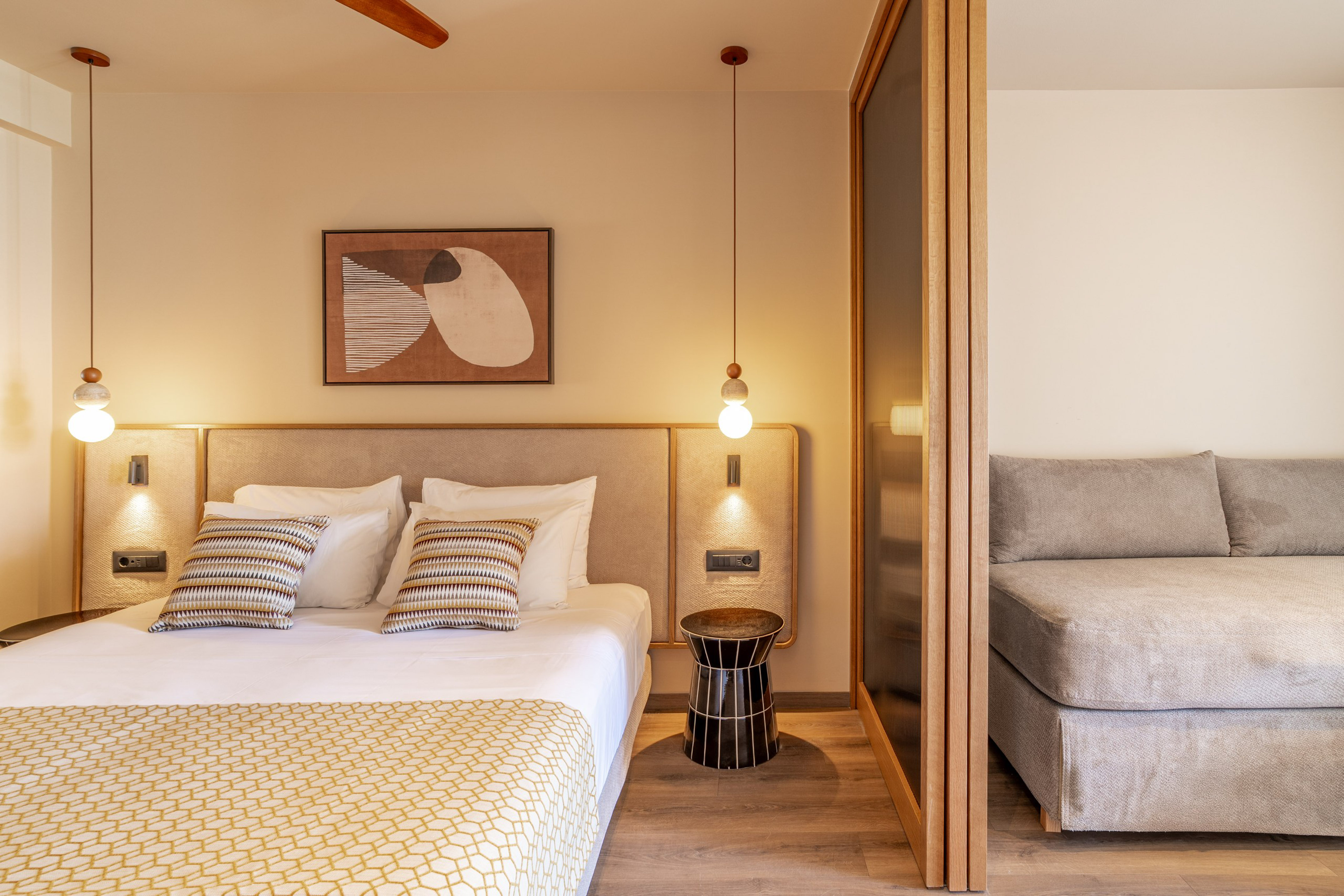 Modern hotel bedroom with large bed, abstract art, pendant light, and gray sofa.