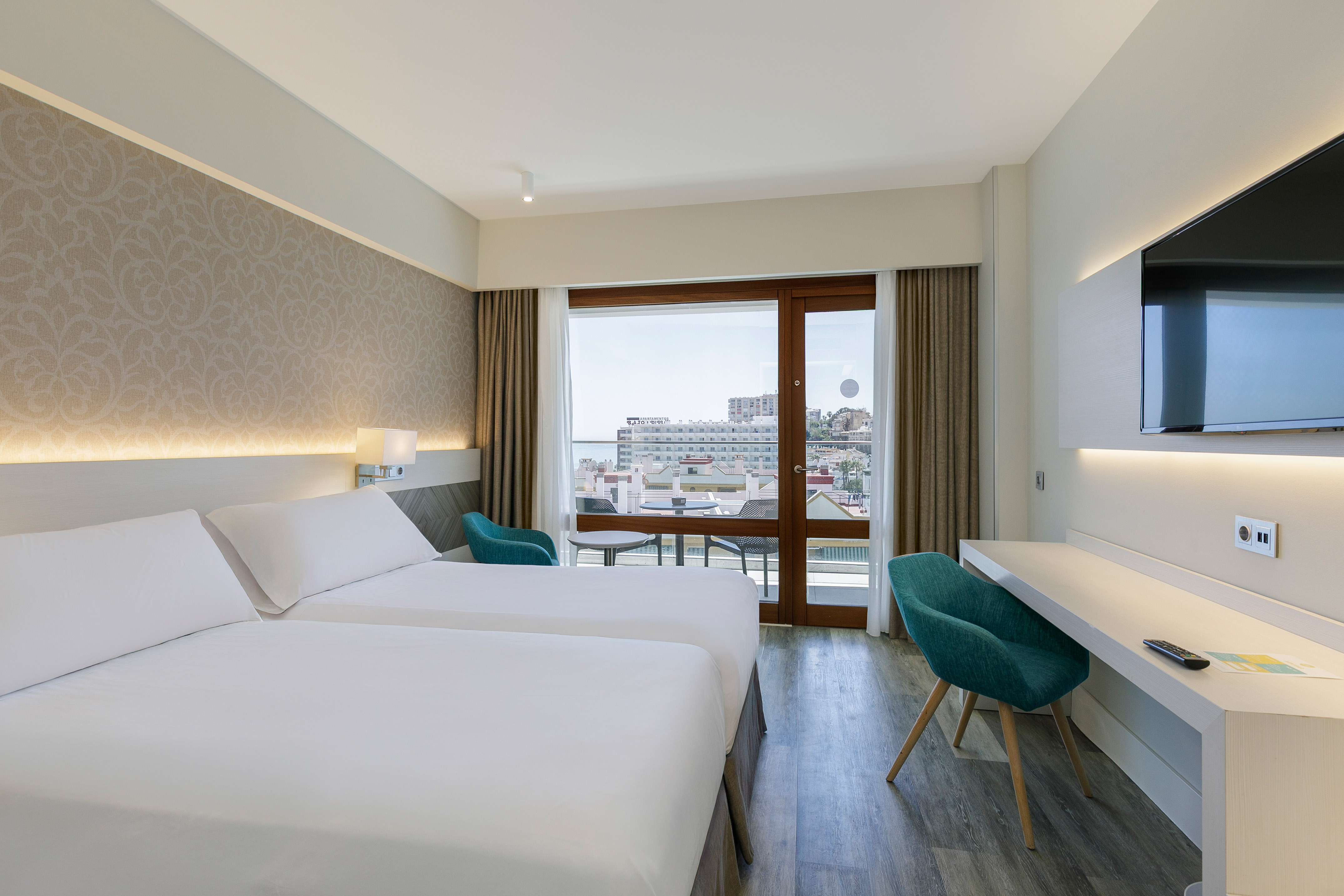 Modern hotel room with twin beds, desk, TV, and balcony with city/sea view.