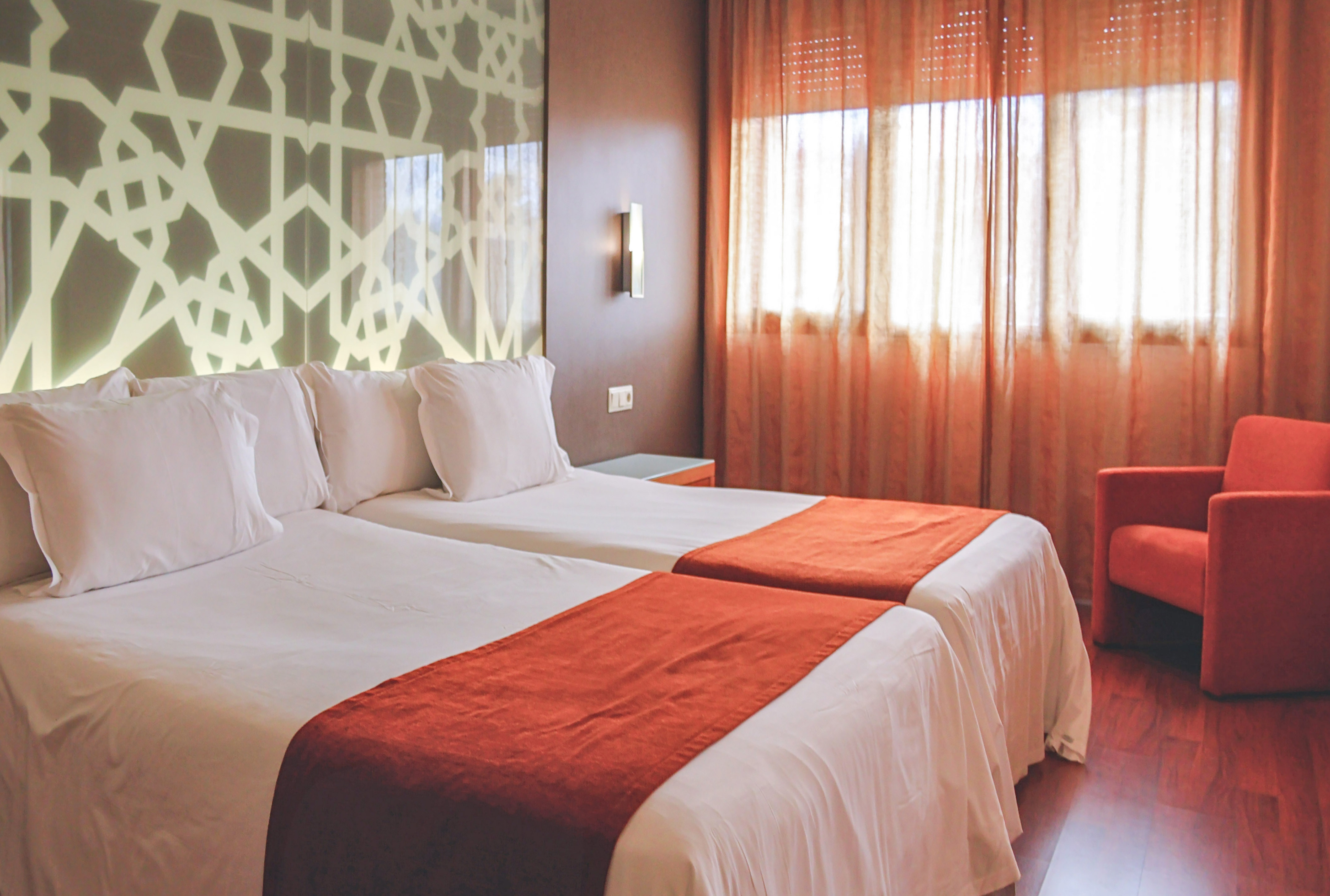 Hotel room with twin beds, white linens, orange accents, and geometric headboard.