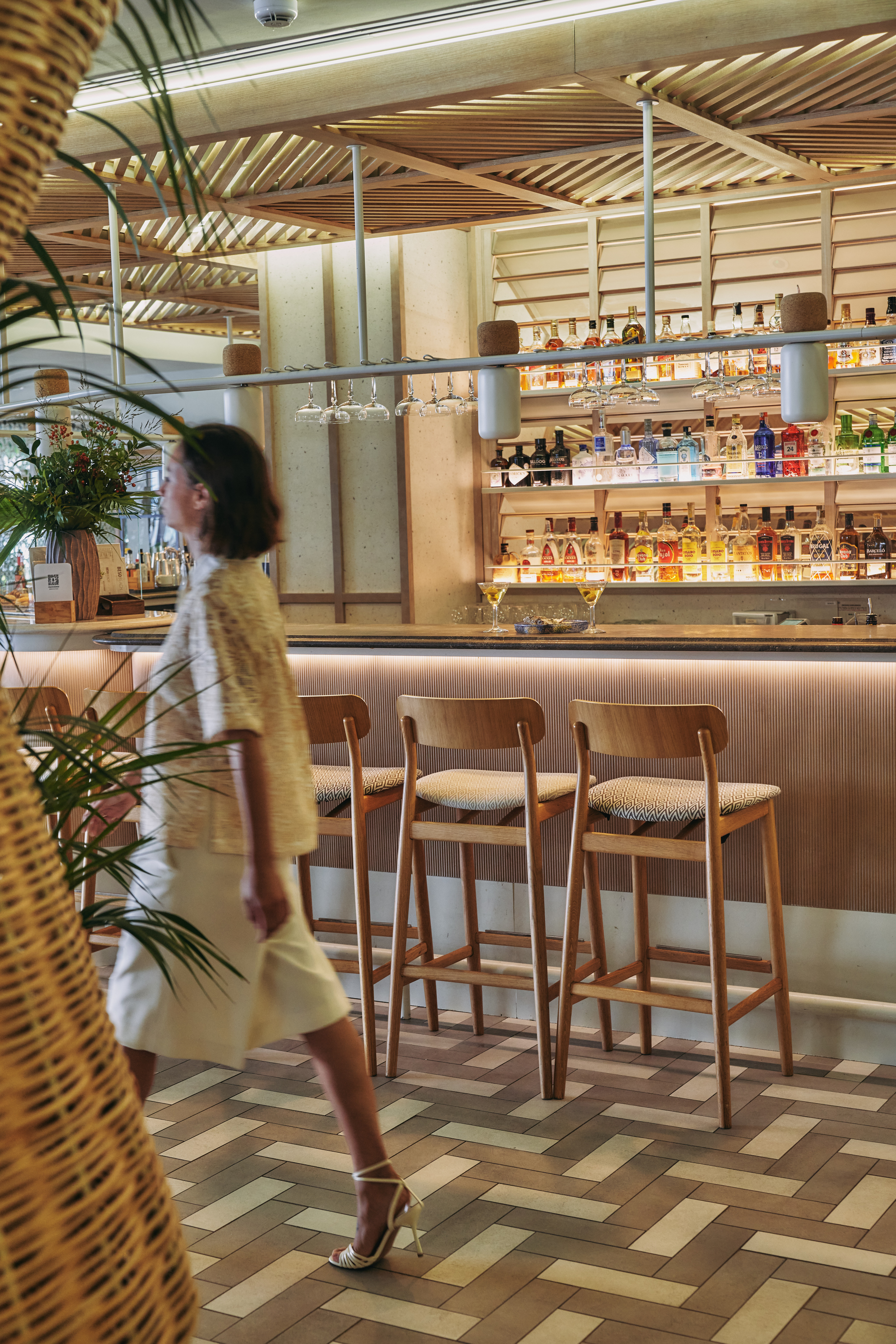 Woman in modern bar interior with liquor display, wooden stools, and chic decor.