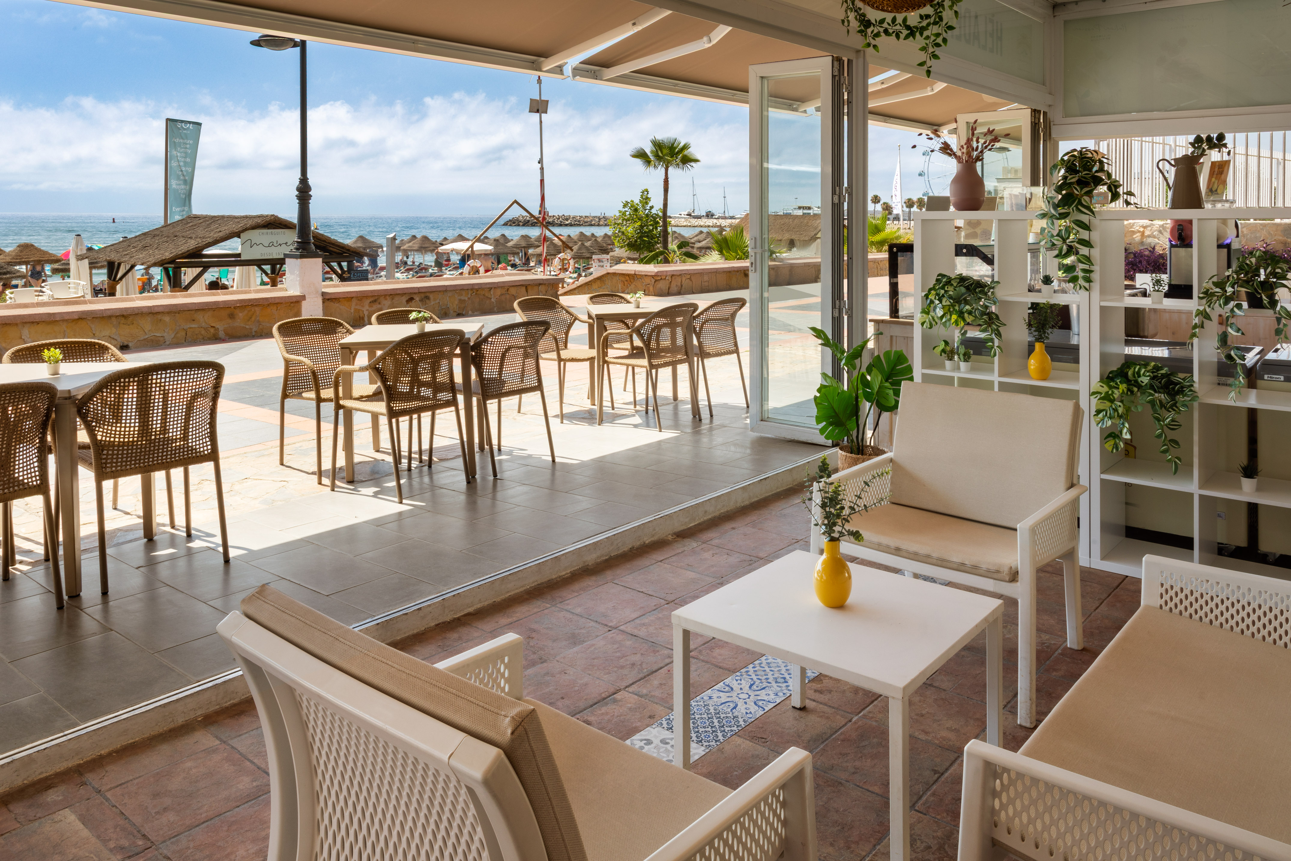 Terraza de chiringuito en la playa con mesas, sillas y vista al mar.