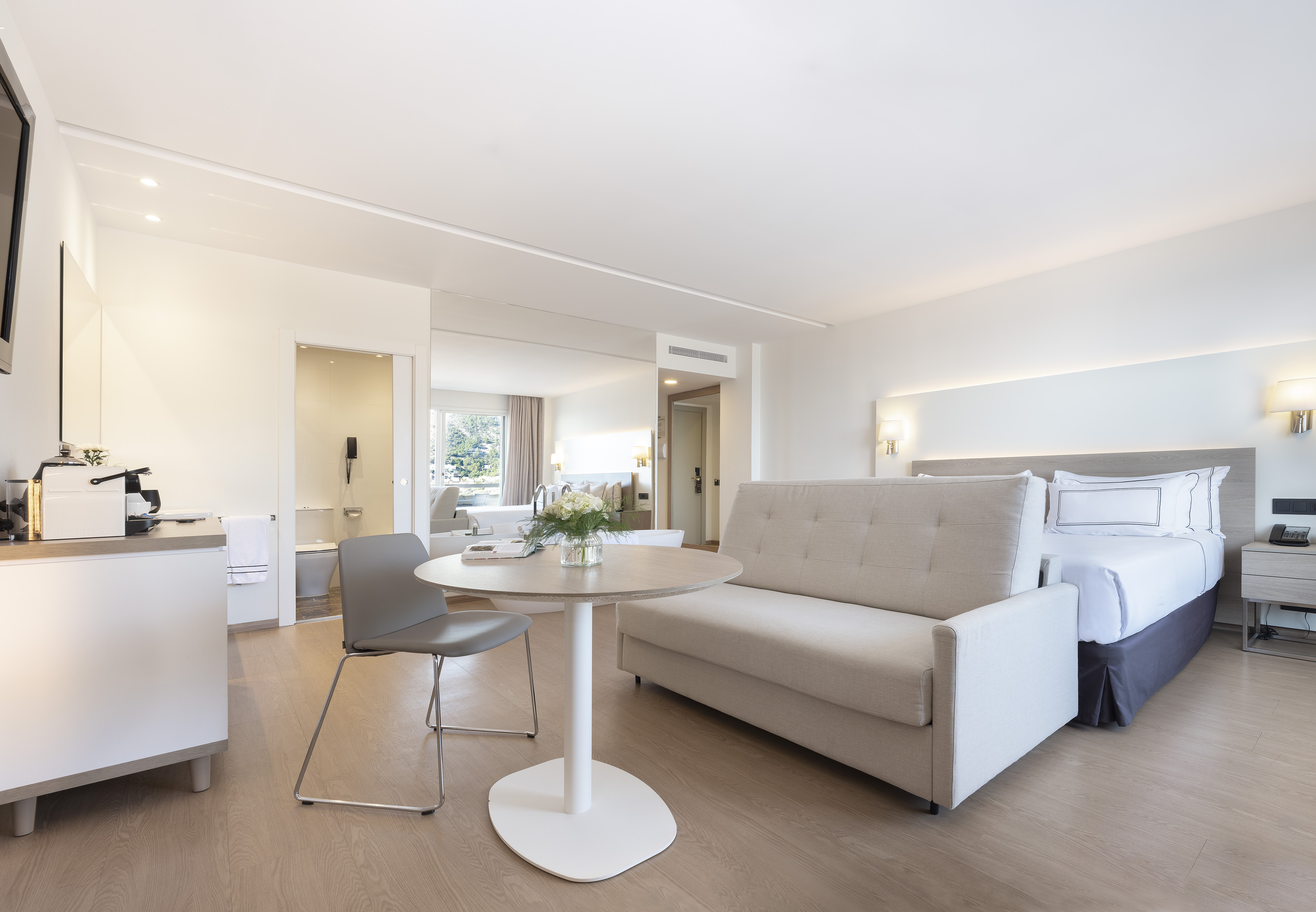 Bright modern studio apartment with bed, sofa, small table, and warm wood flooring.