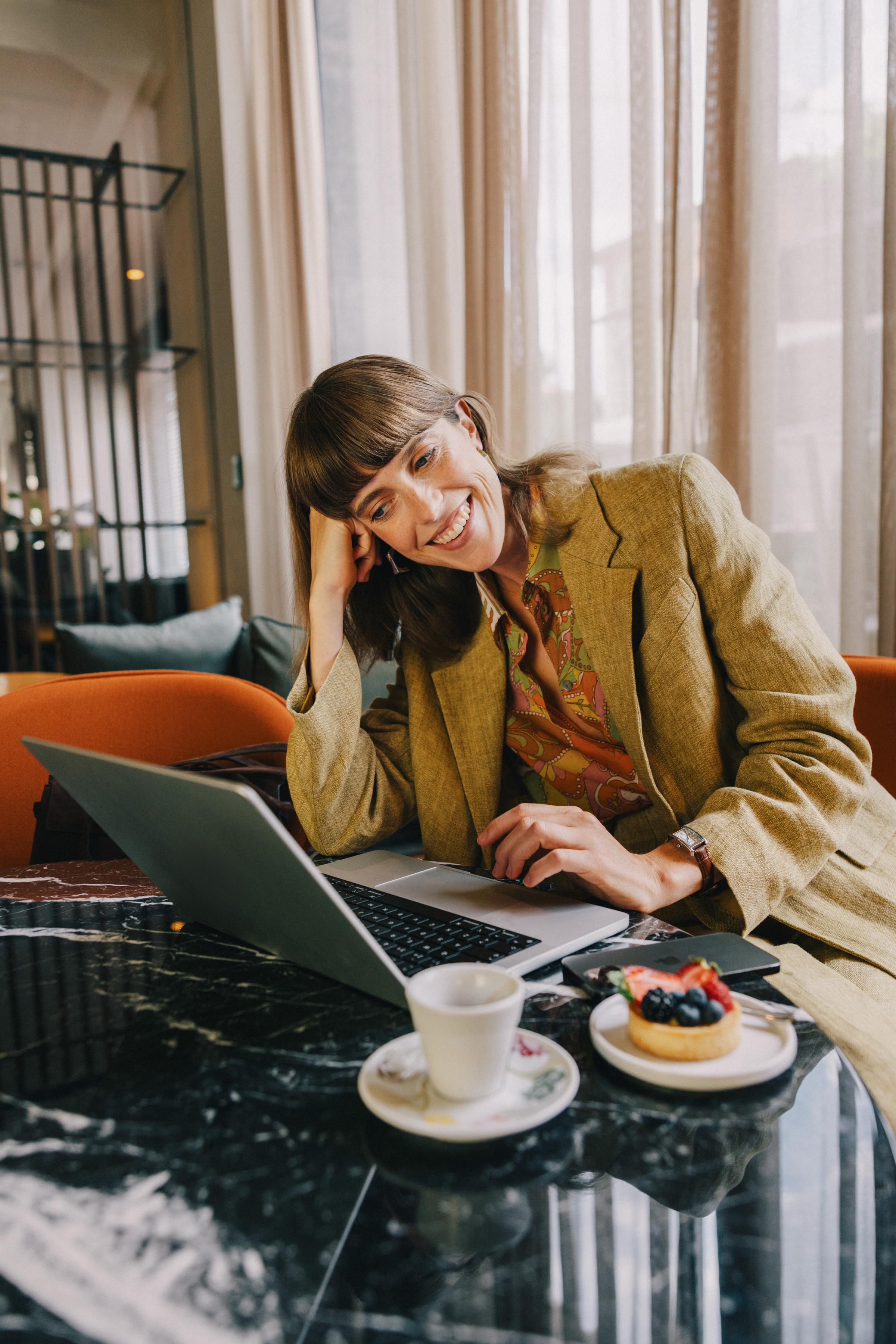 Smiling woman working on laptop at cafe table with coffee and berry tart.