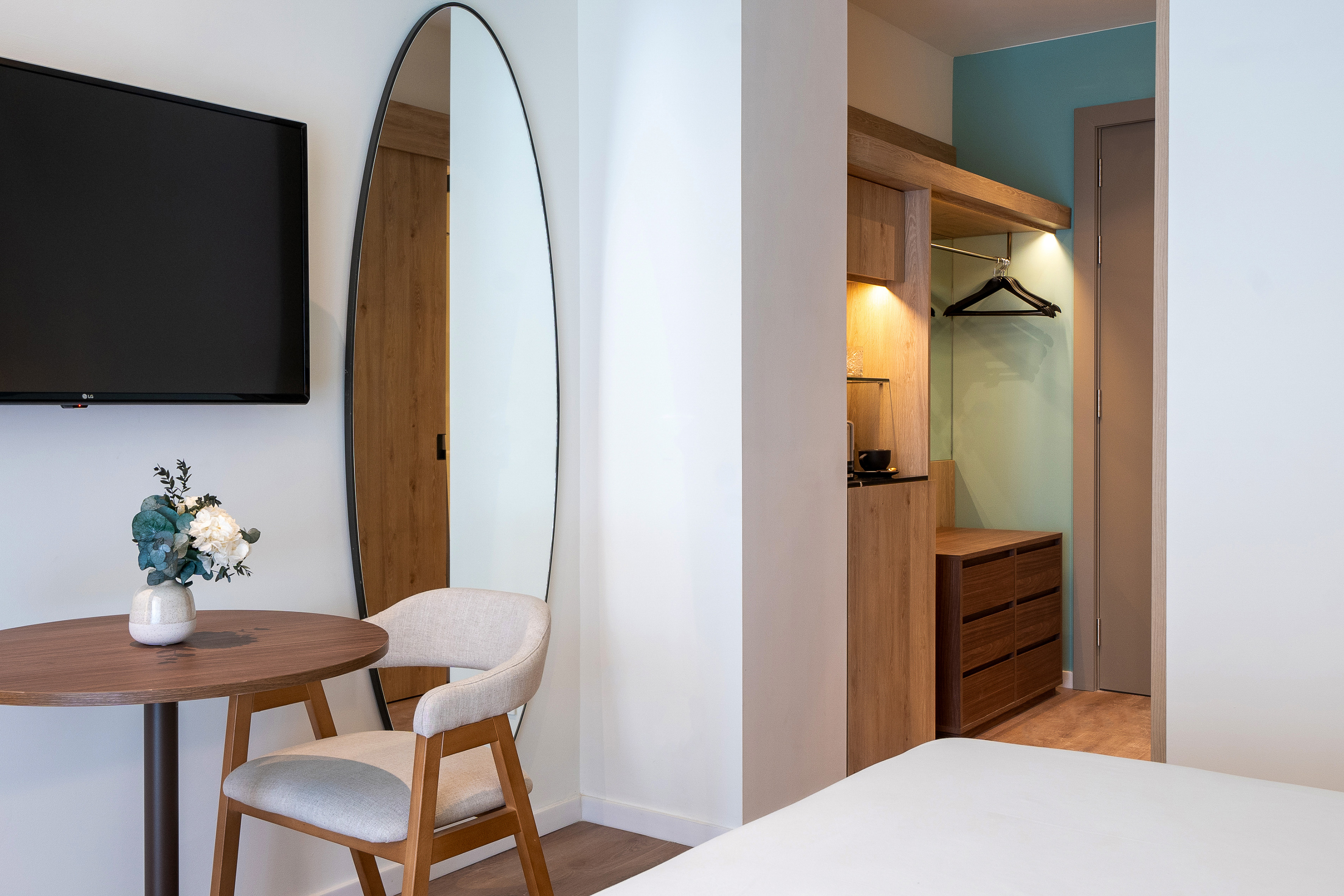 Modern hotel bedroom with TV, oval mirror, table, and open closet.
