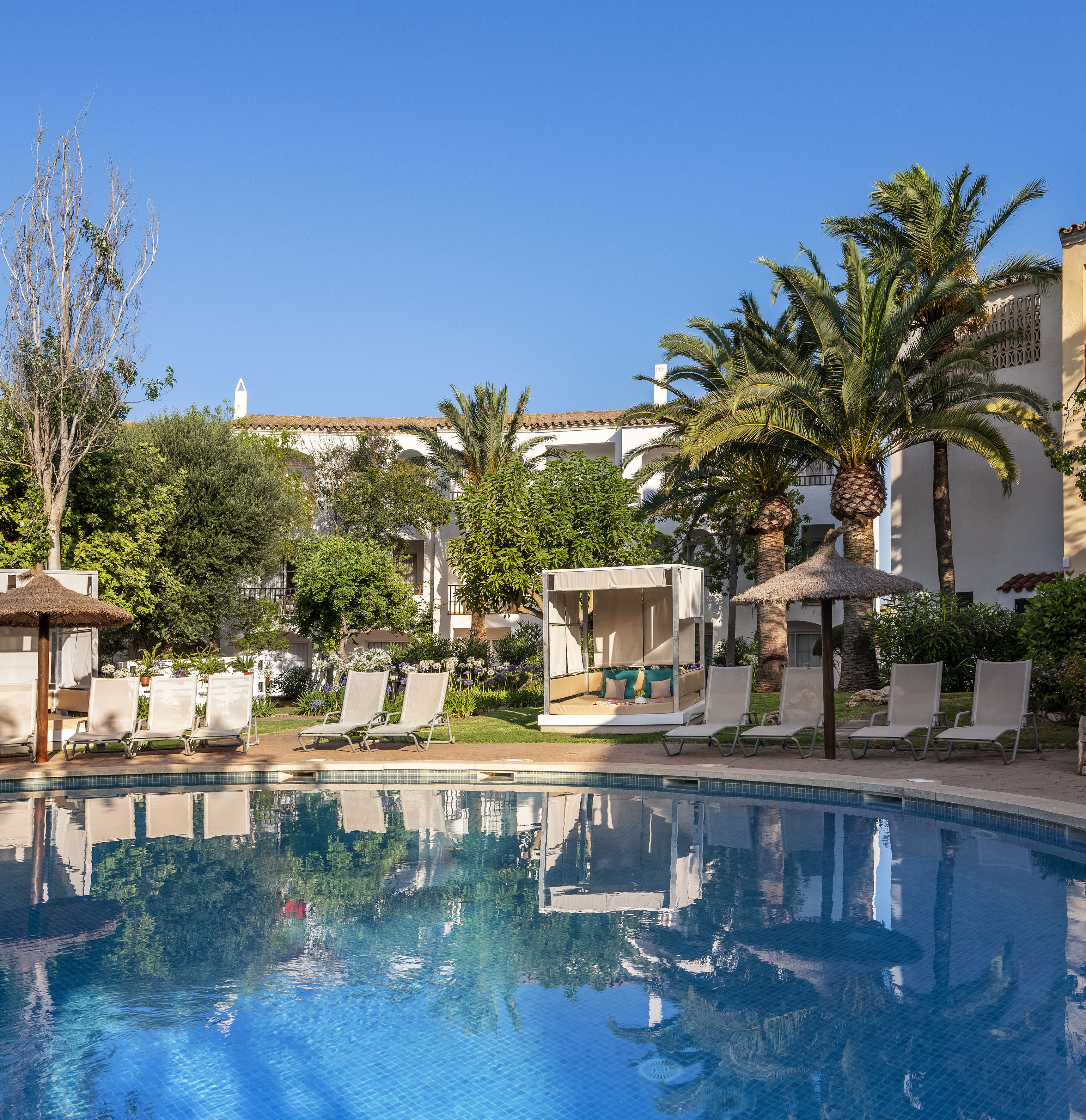 Mediterranean resort pool with sun loungers, cabana, and palm trees.