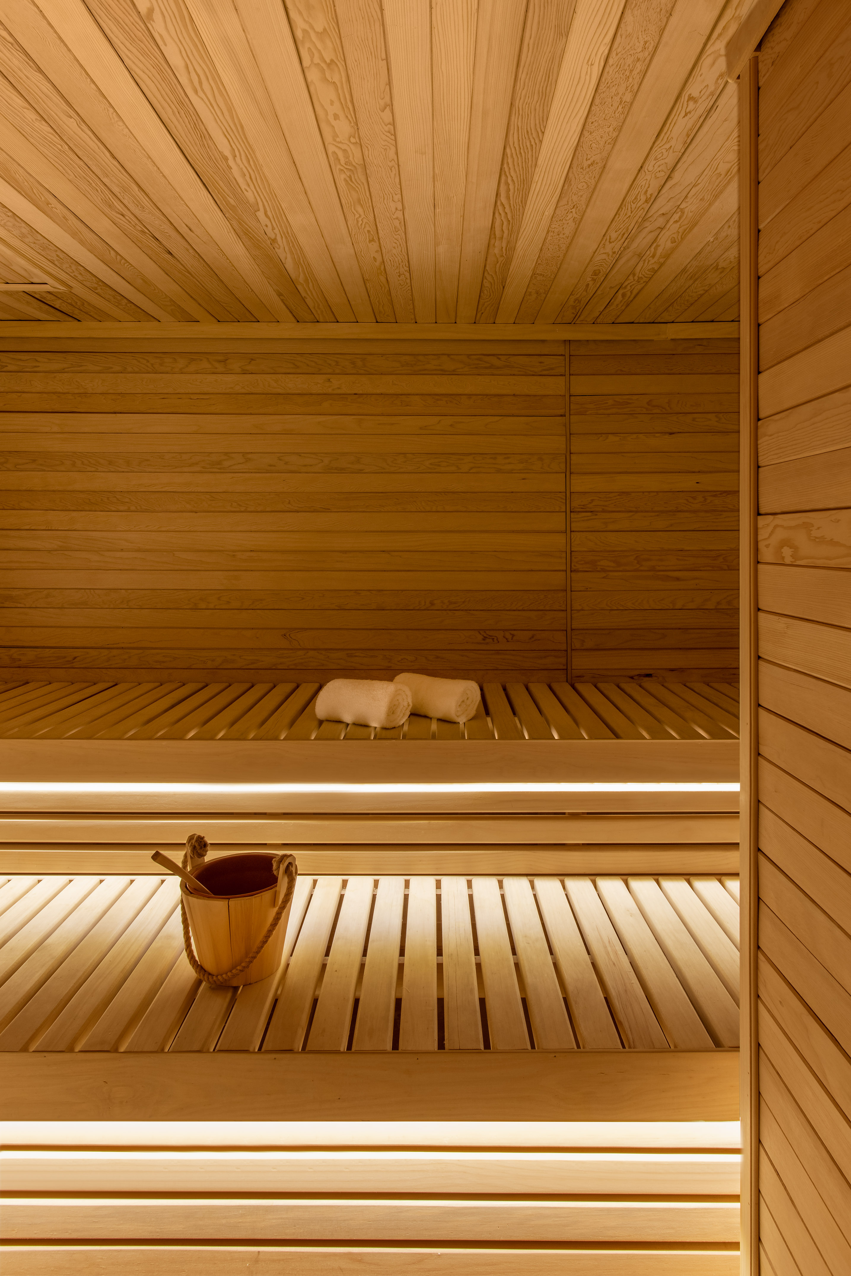 Sauna with light wooden benches, soft lighting, rolled towels, and a wooden bucket.