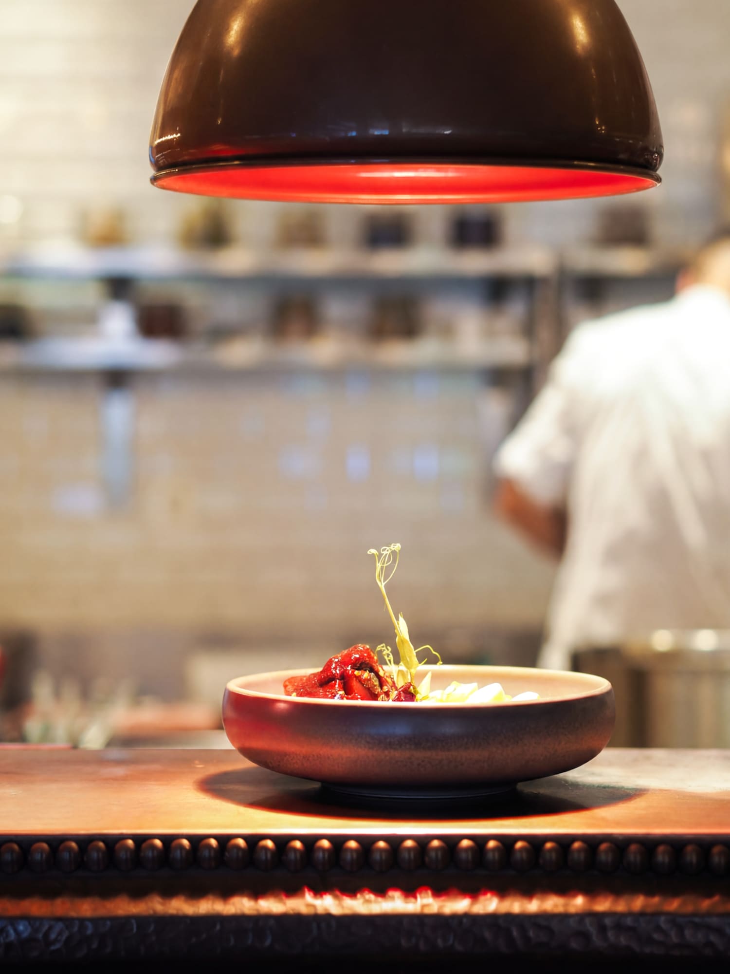 Gourmet dish under red heat lamp on kitchen counter, chef in background.