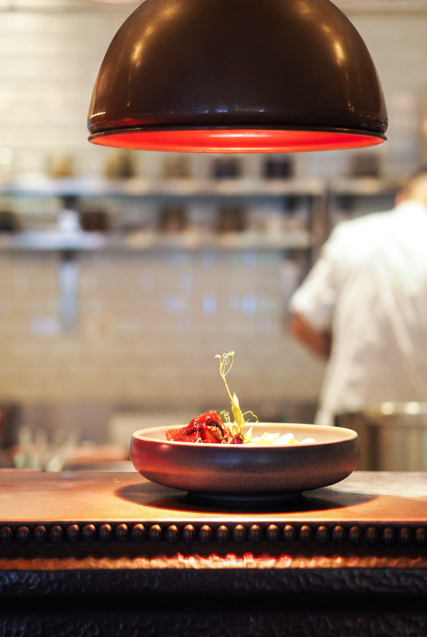 Gourmet dish under red heat lamp on kitchen counter, chef in background.