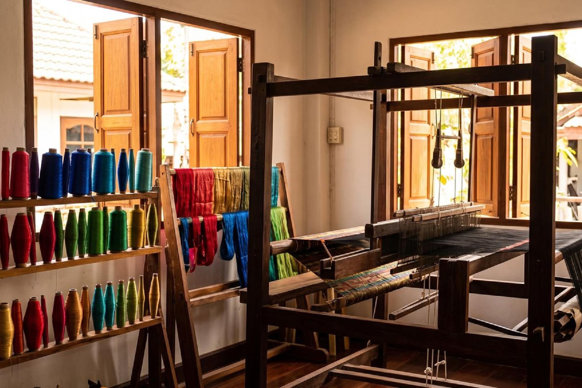 Wooden weaving loom with shelves of colorful yarn spools in a sunlit craft room.