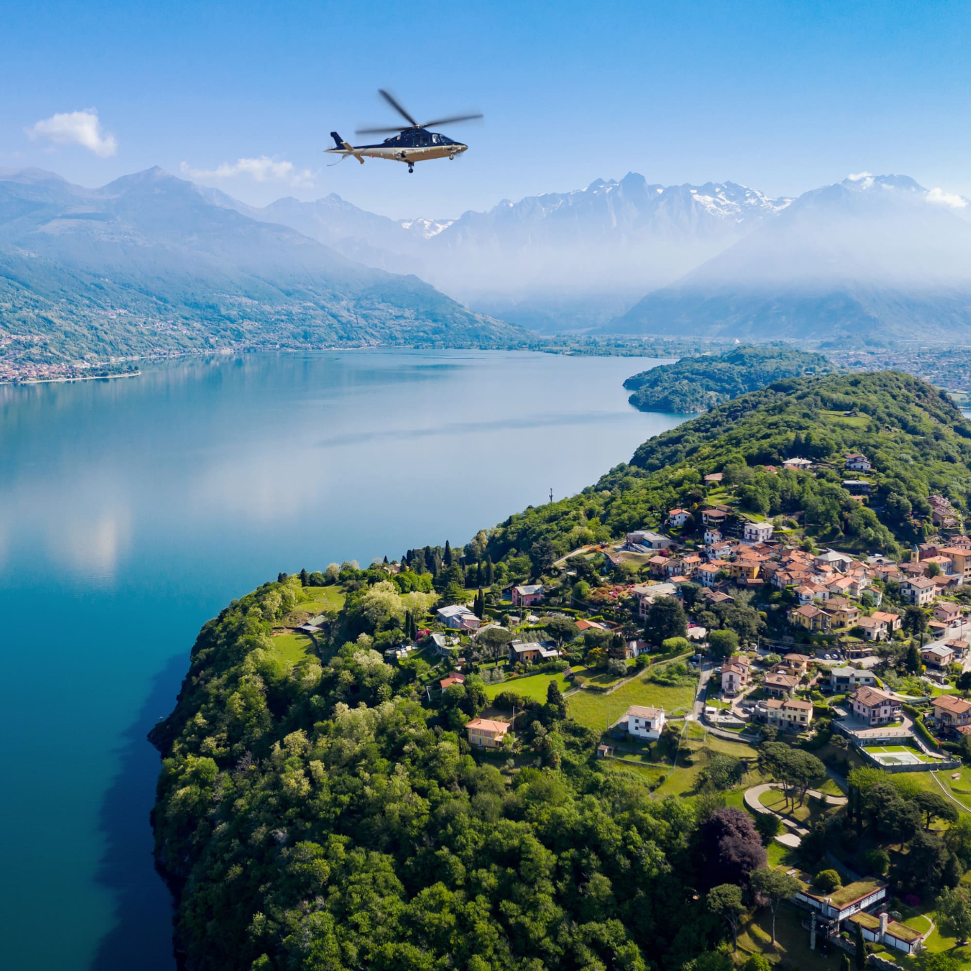 Helicopter above scenic Lake Como village, green peninsula, and misty mountains.