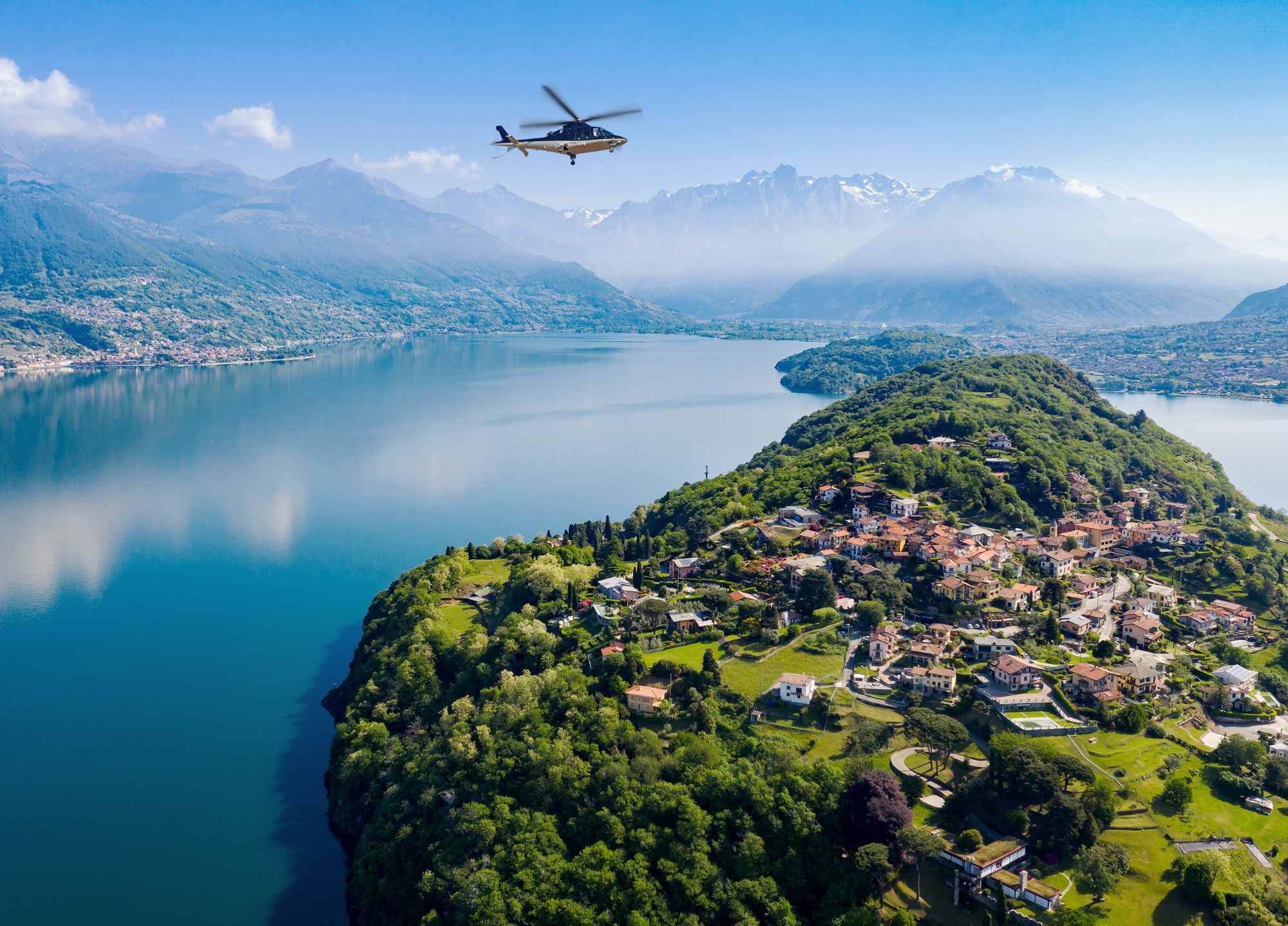 Helicopter above scenic Lake Como village, green peninsula, and misty mountains.