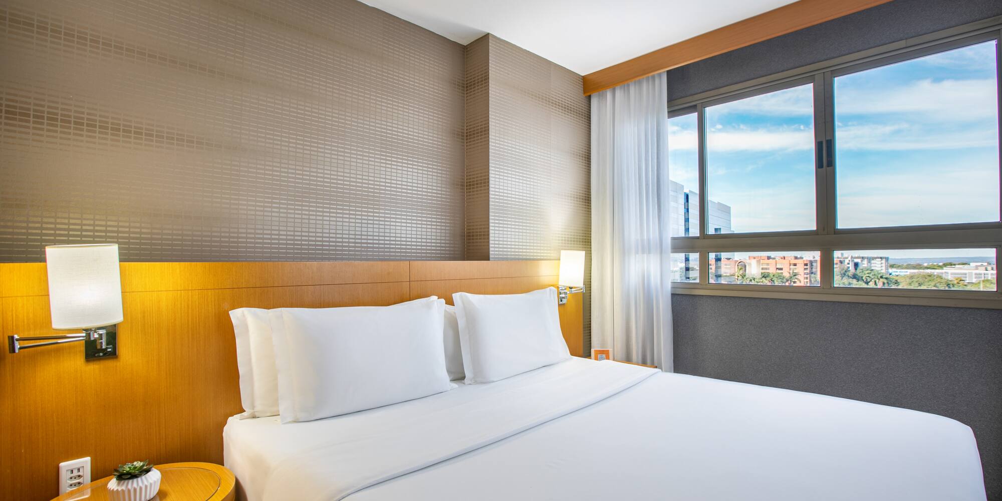 Bright modern hotel room with white bed, wooden headboard, and city view.