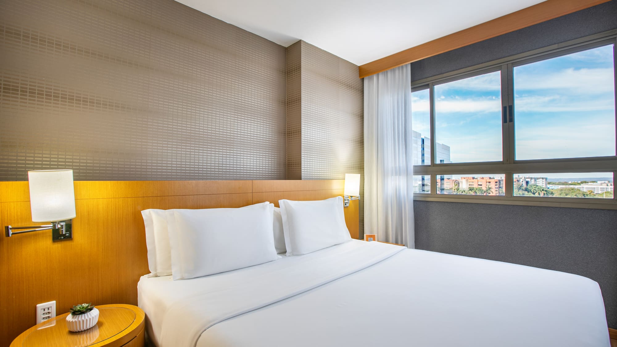 Bright modern hotel room with white bed, wooden headboard, and city view.