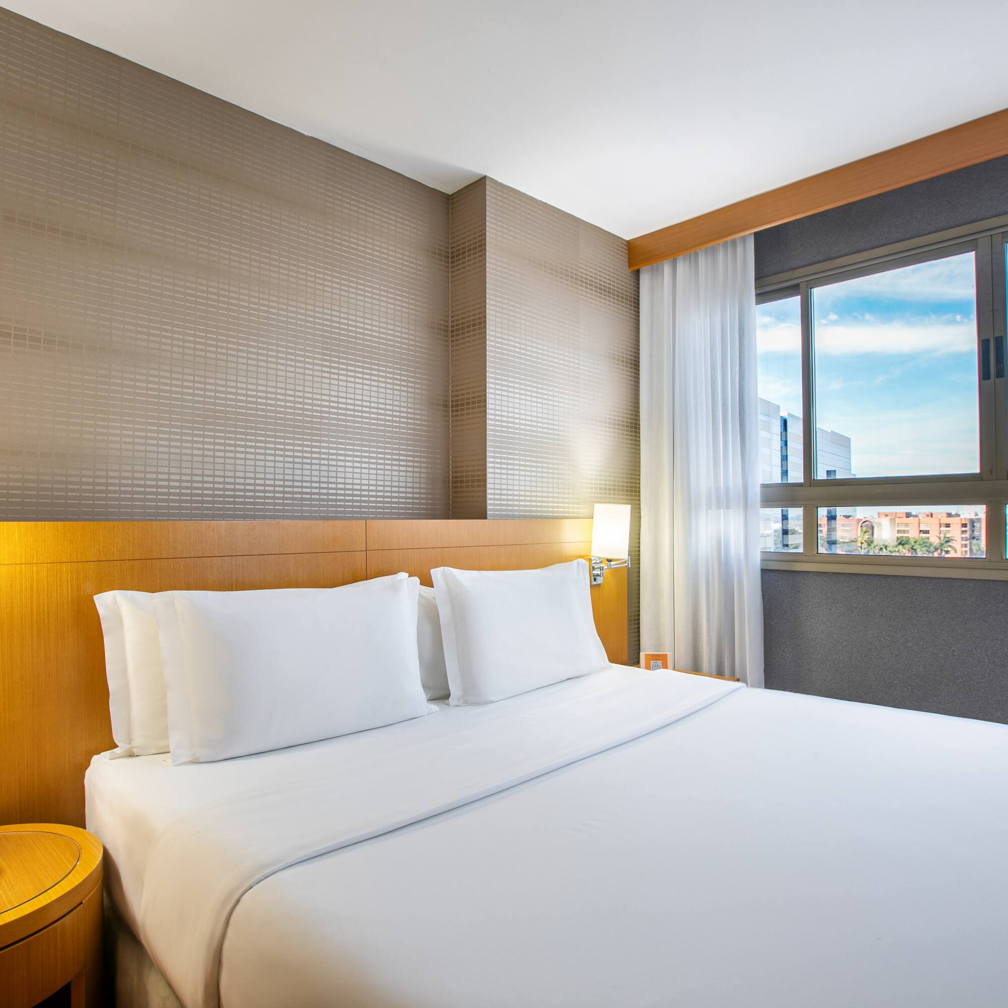 Bright modern hotel room with white bed, wooden headboard, and city view.