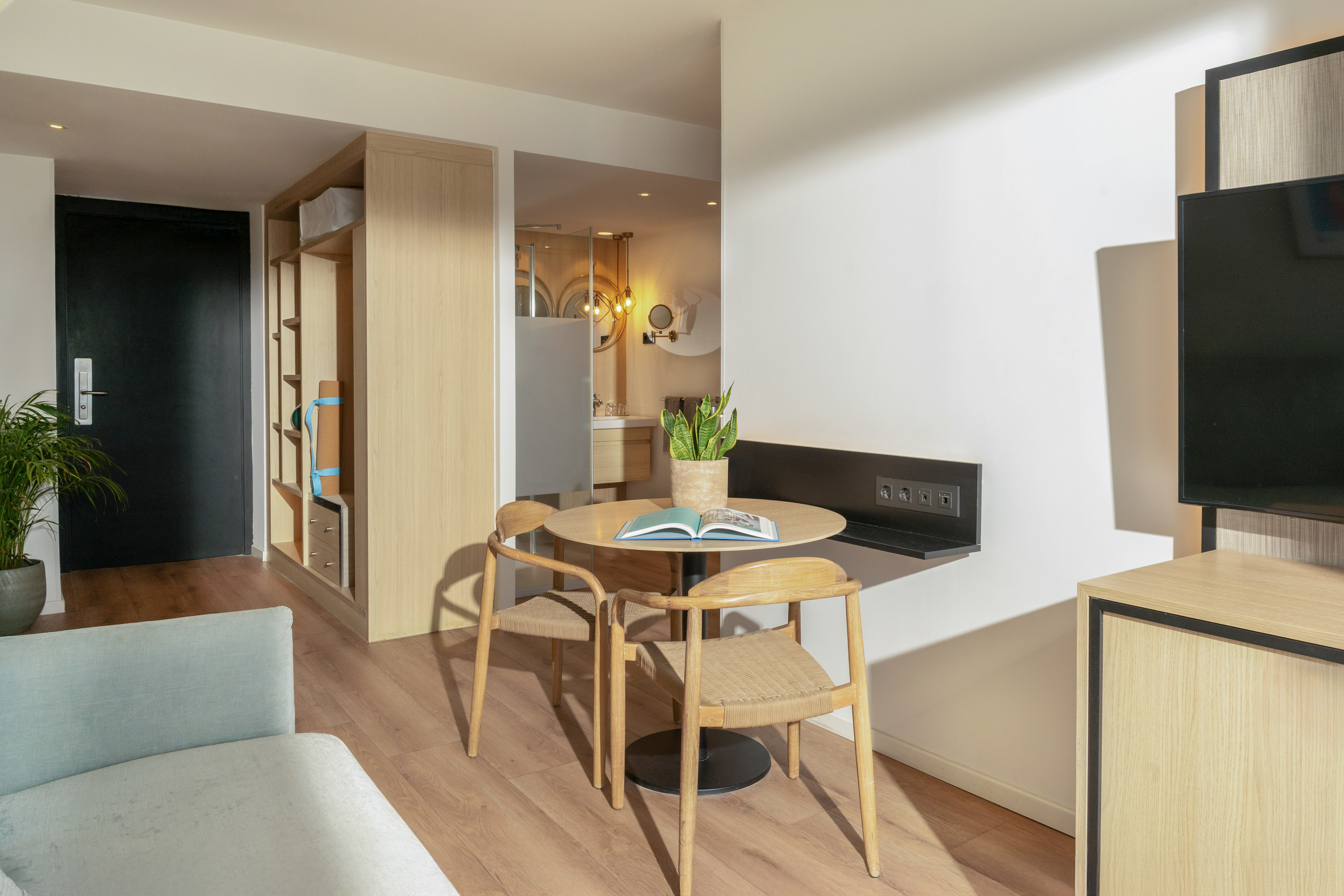 Modern hotel suite interior with wooden dining area, chairs, and light sofa.
