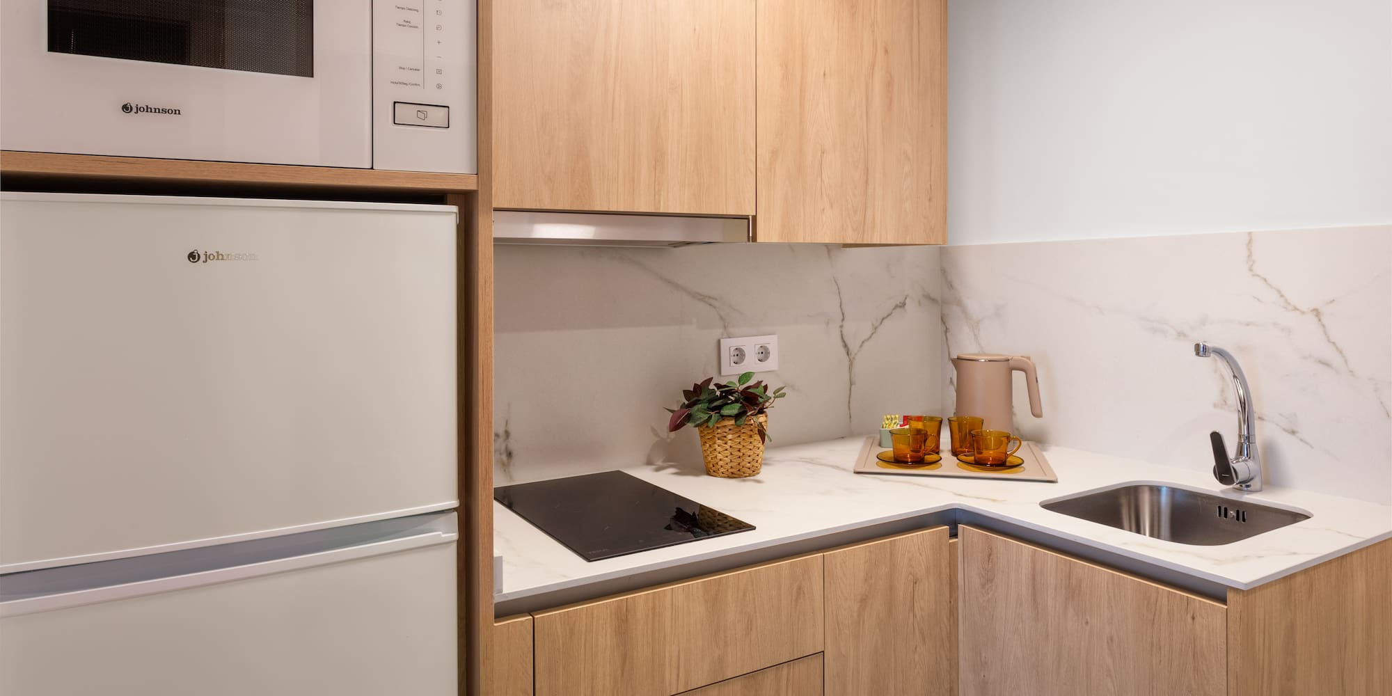 Compact modern kitchen with light wood cabinets, marble counter, microwave, fridge, sink.
