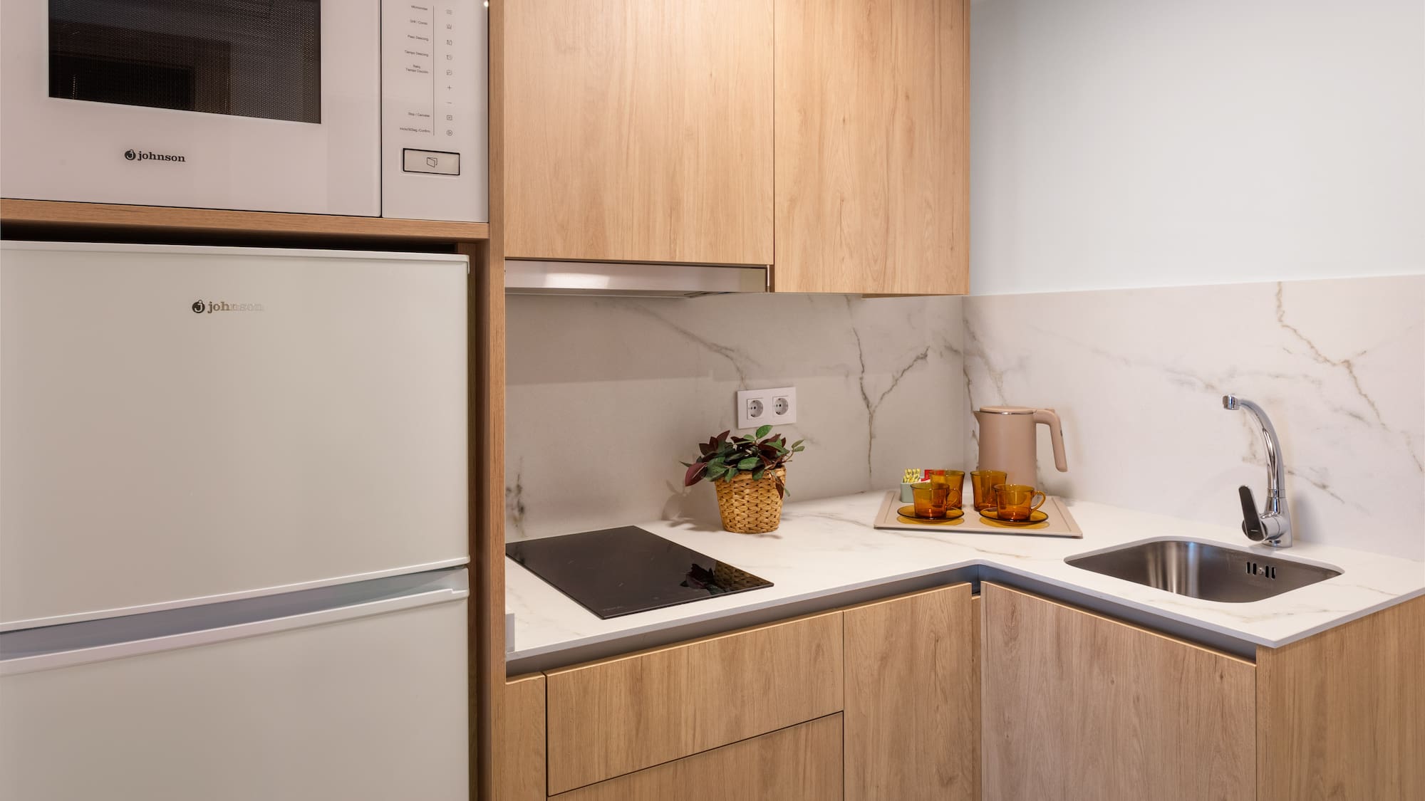 Compact modern kitchen with light wood cabinets, marble counter, microwave, fridge, sink.