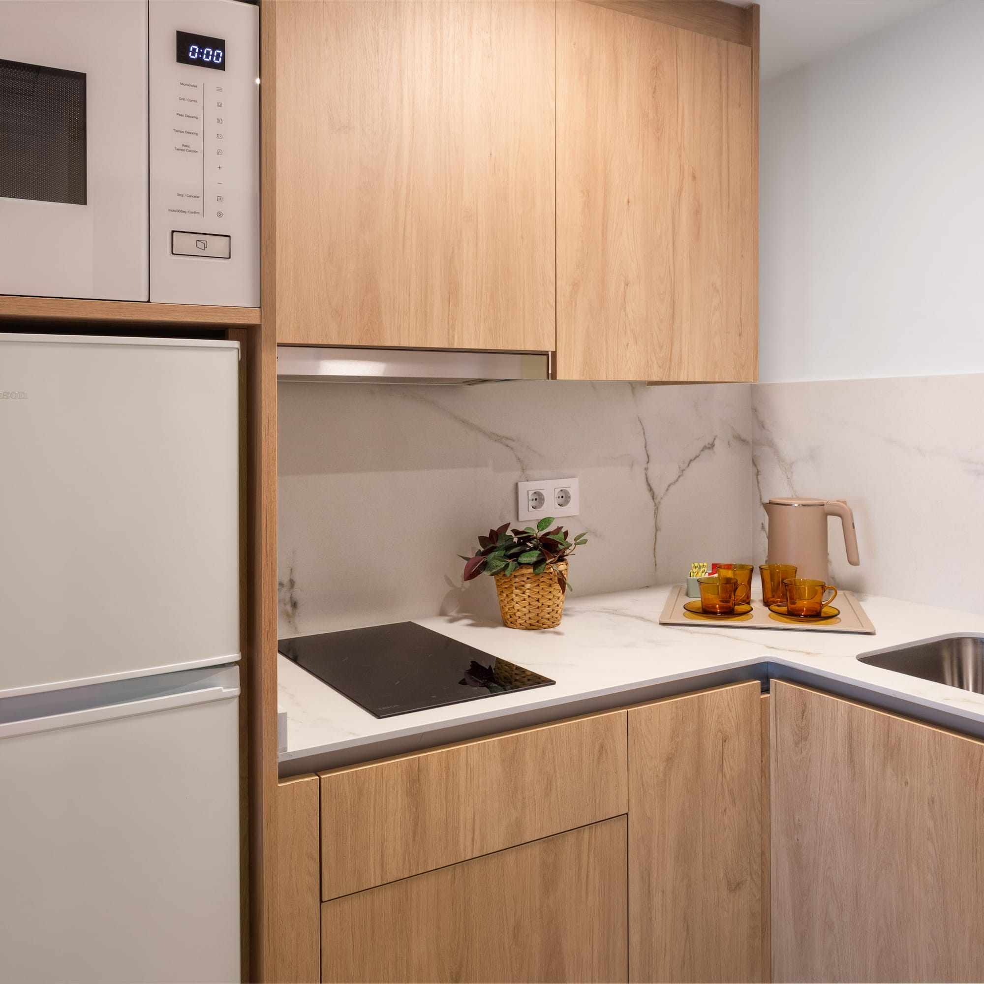 Compact modern kitchen with light wood cabinets, marble counter, microwave, fridge, sink.