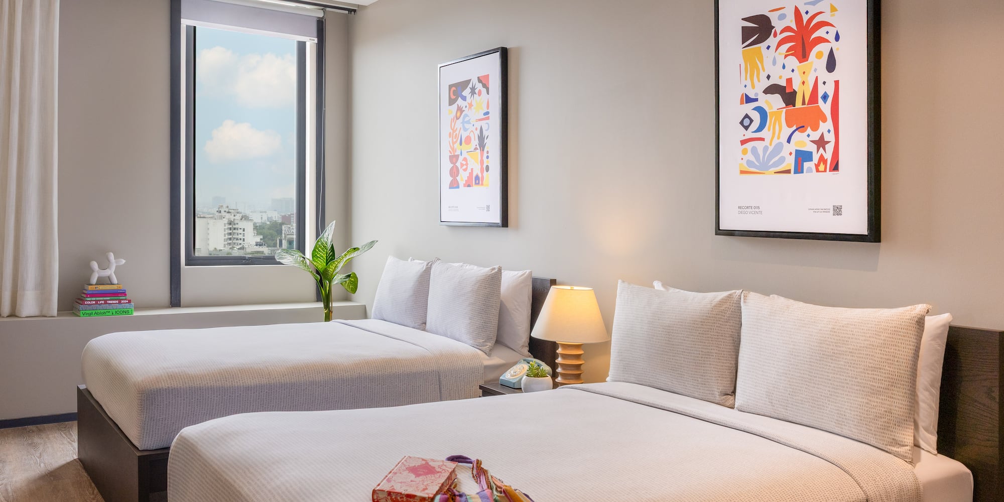 Contemporary hotel room with twin beds, city view window, and vibrant artwork.