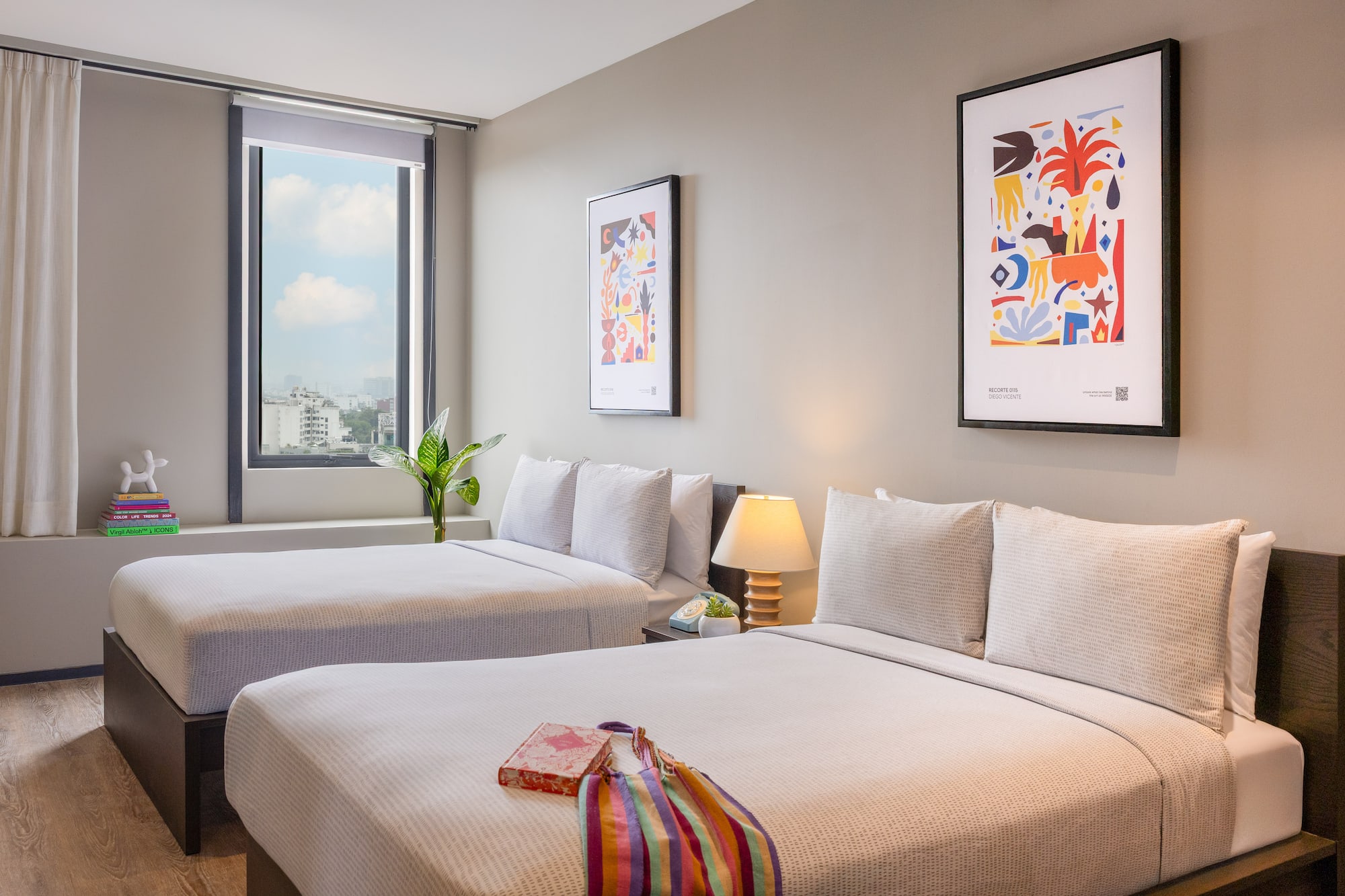 Contemporary hotel room with twin beds, city view window, and vibrant artwork.