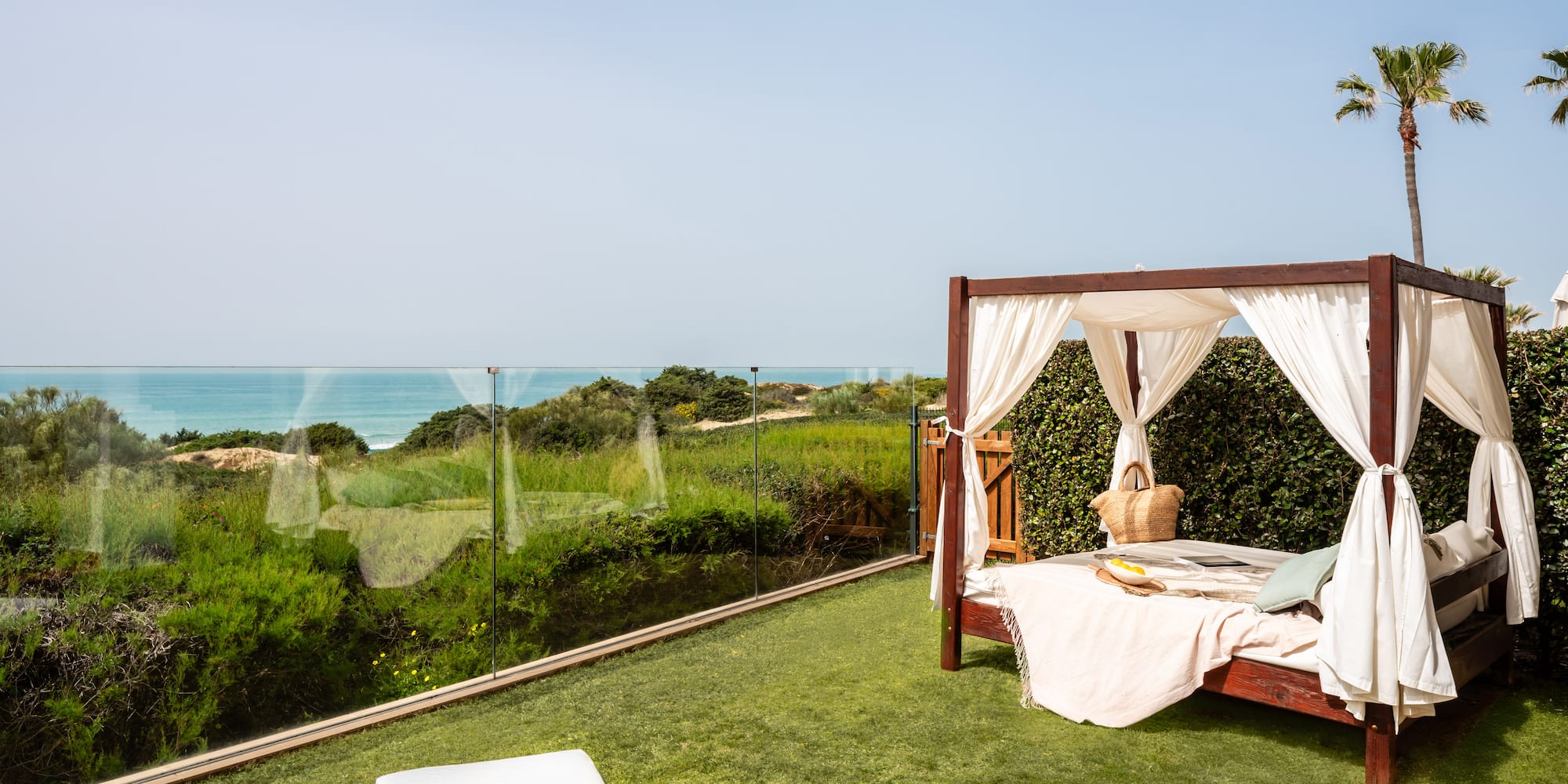 Outdoor canopy bed on green lawn with ocean view and palm trees.