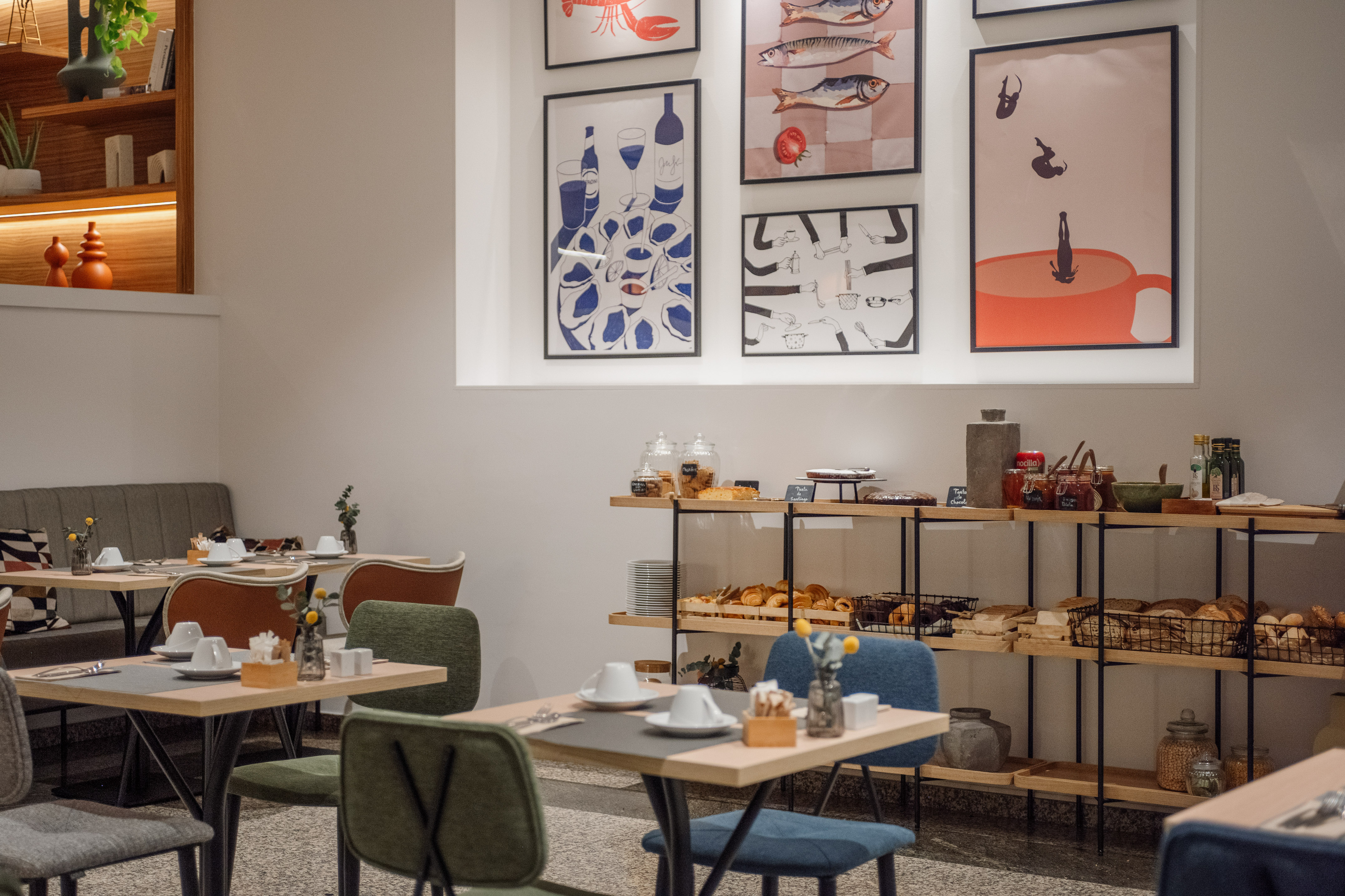 Modern hotel breakfast room with buffet, pastries, set tables, and wall art.