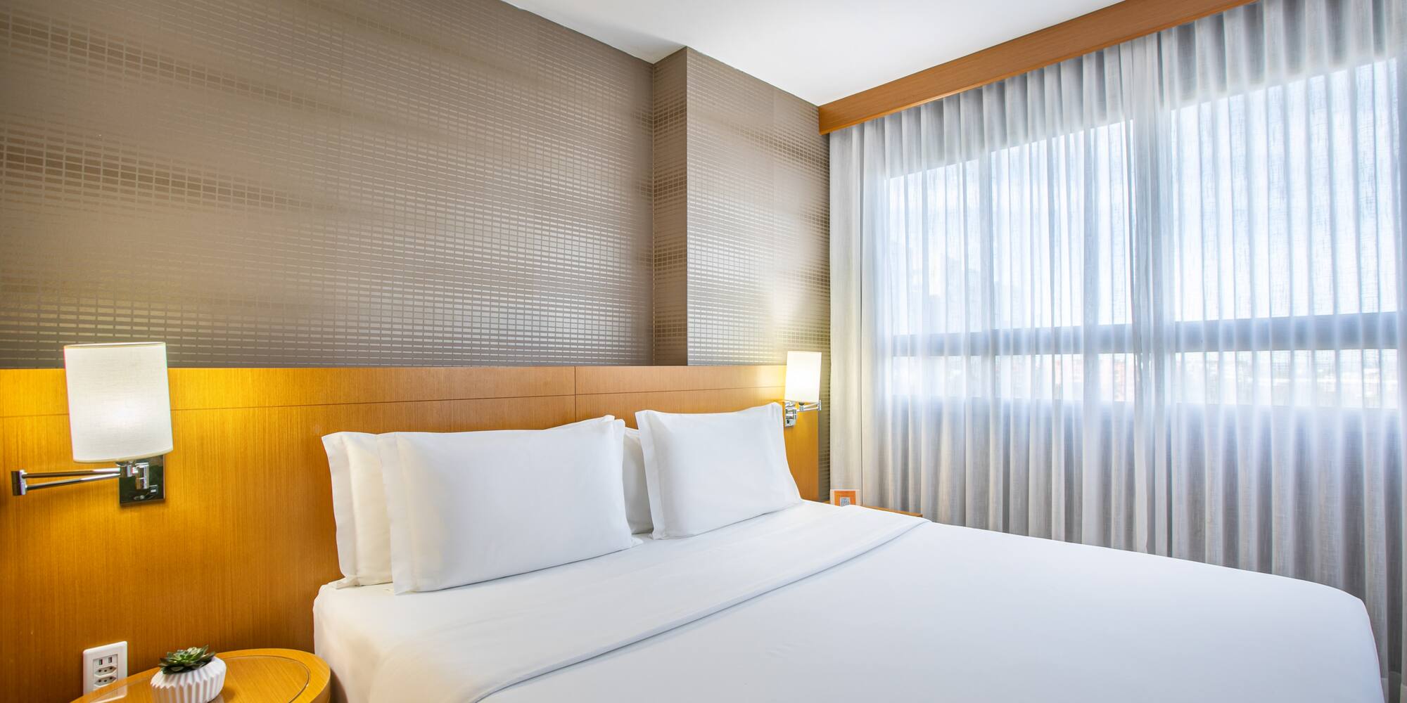 Modern hotel bedroom with white queen bed, wood headboard, and sheer curtains.