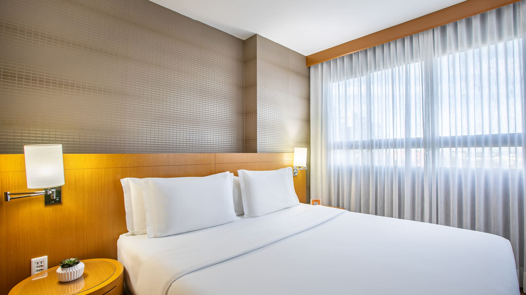 Modern hotel bedroom with white queen bed, wood headboard, and sheer curtains.