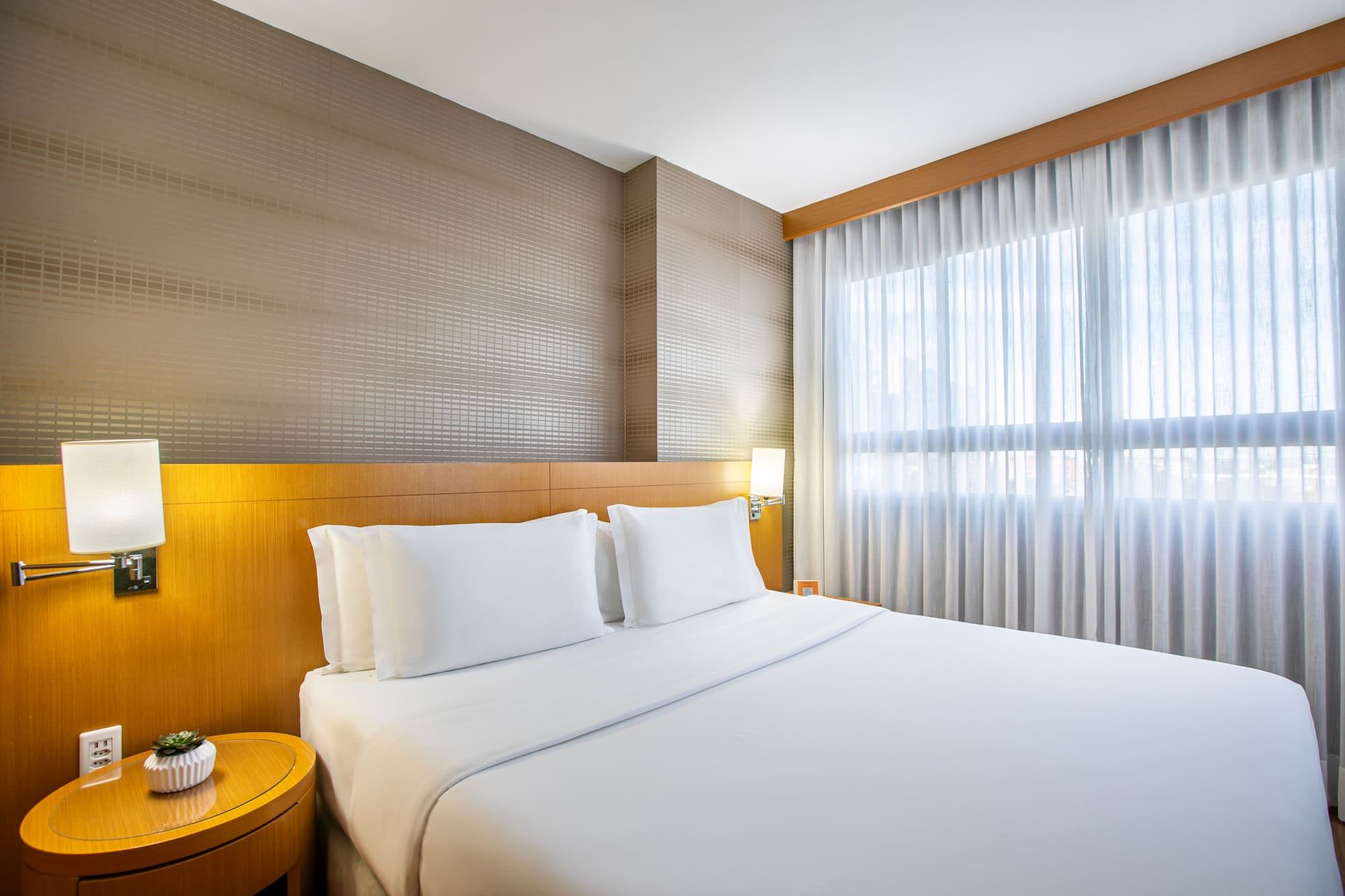 Modern hotel bedroom with white queen bed, wood headboard, and sheer curtains.